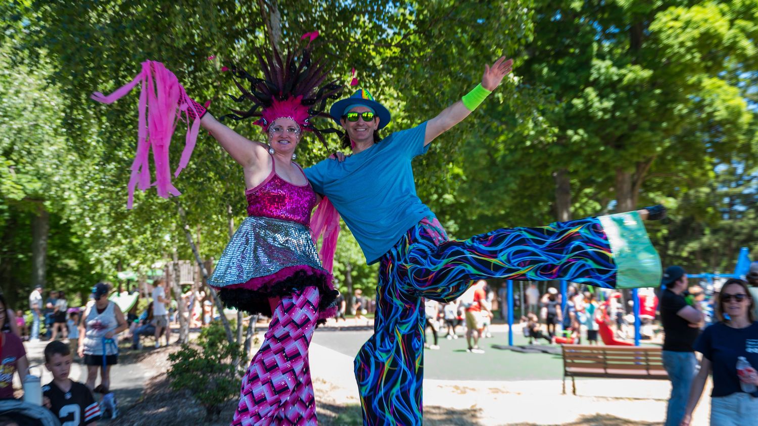 Two colorful stilt walkers in vibrant costumes perform at an outdoor festival surrounded by trees and crowds.