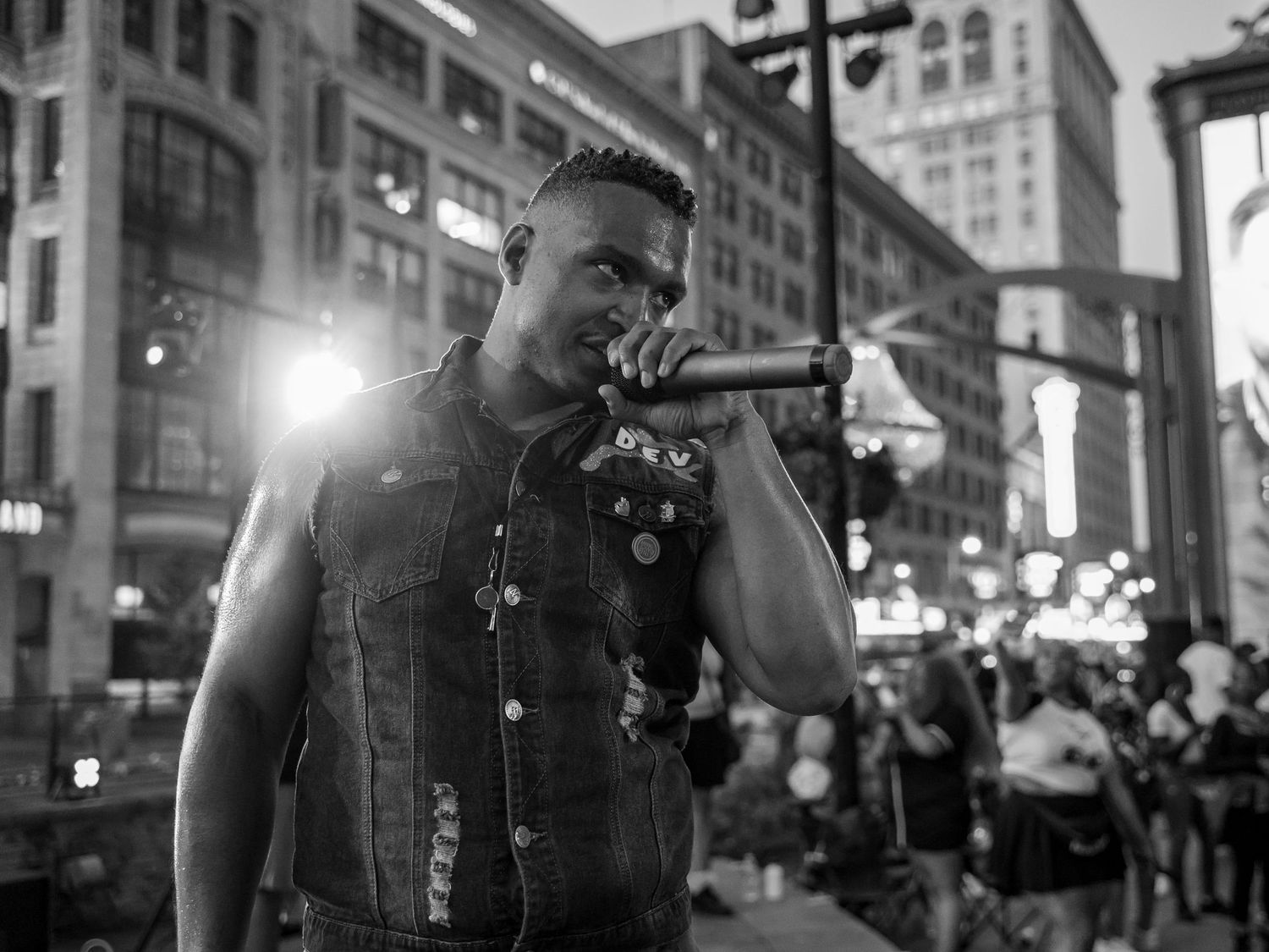 Black and white photo of a man in a denim vest performing with a microphone on a city street at night.