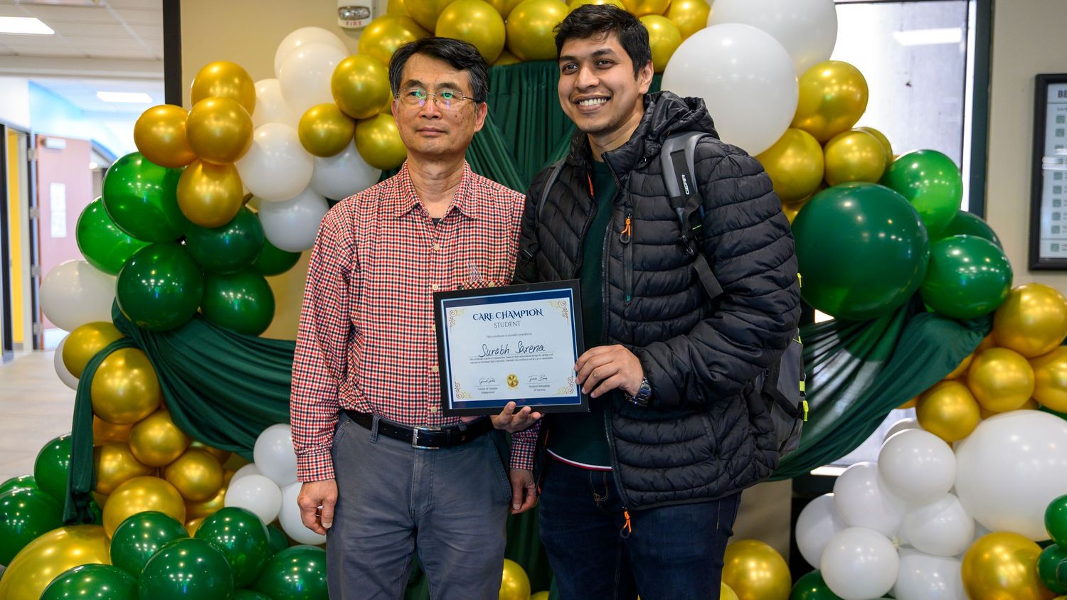 Two men smiling and holding a certificate award in front of green, gold, and white balloon arch decoration.