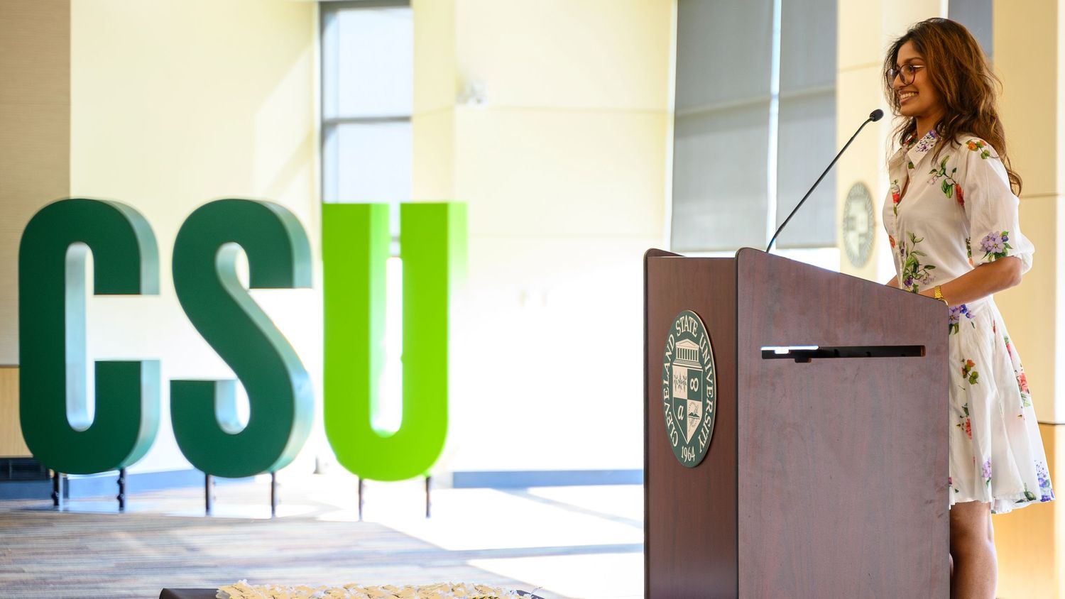 Woman speaking at podium beside large CSU letters sign at California State University event.