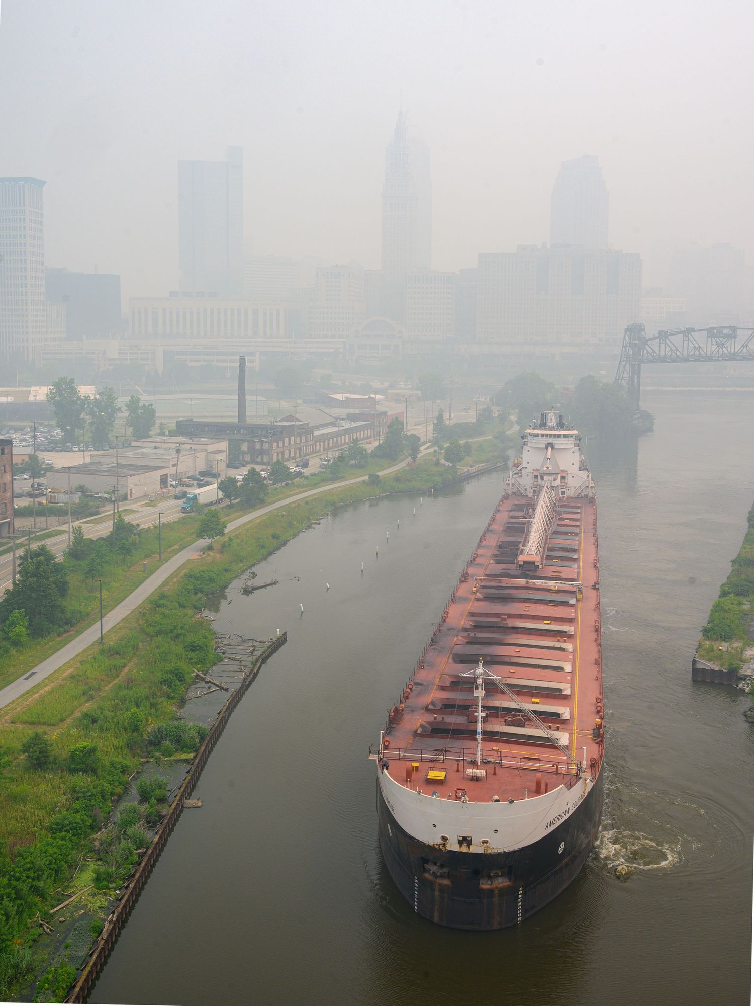 Large cargo ship navigating a narrow river through an industrial city on a hazy, smoggy day.