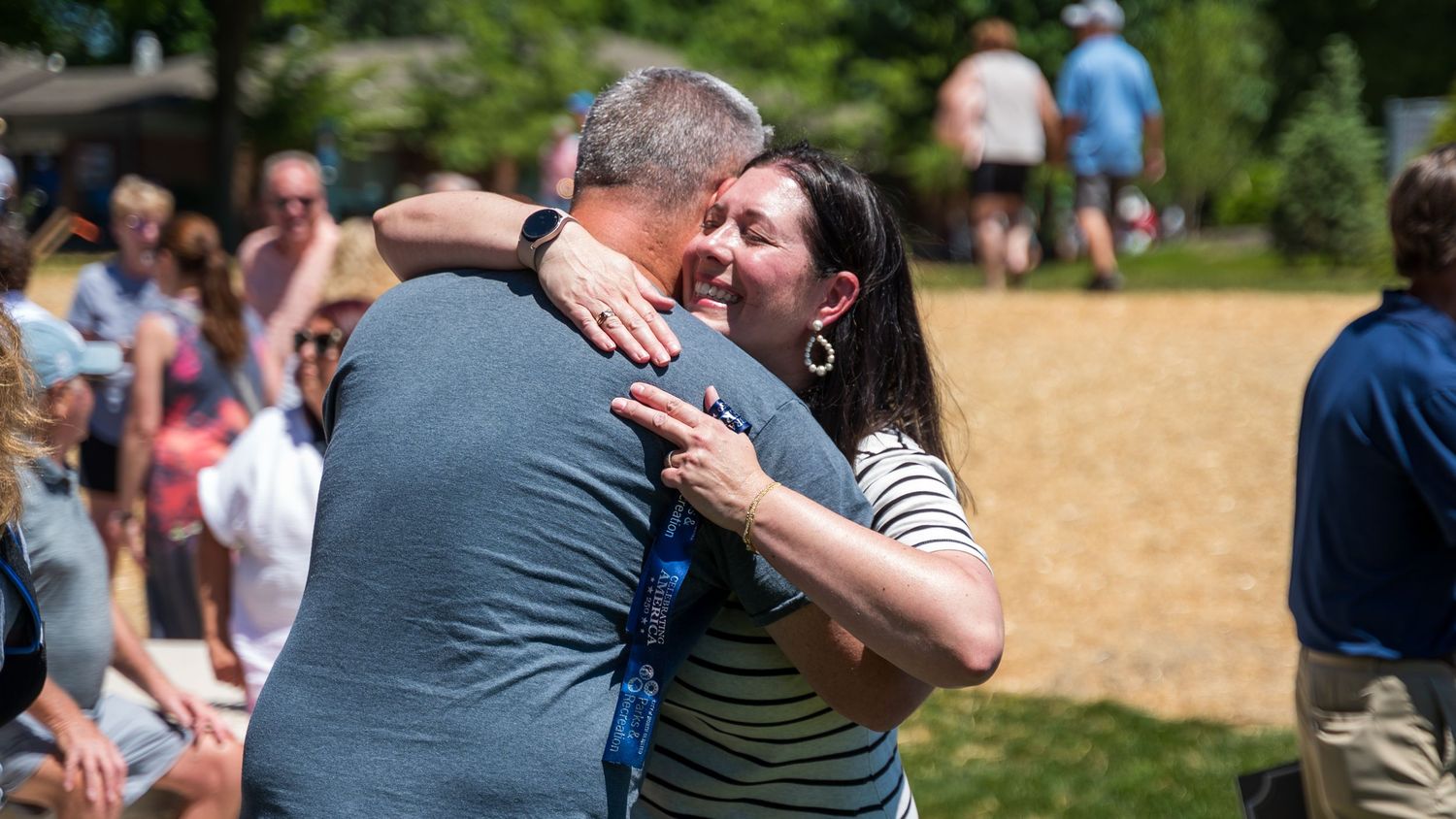 Two people share a warm hug outdoors at a sunny park gathering with trees in the background.