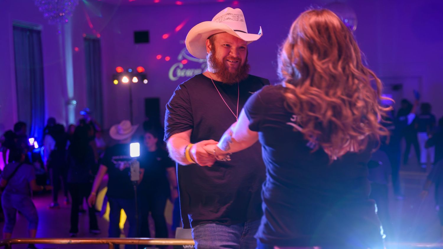 Couple dancing together at a vibrant purple-lit line dance event with cowboy hats.