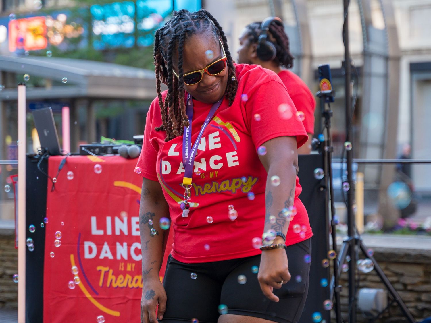 Woman in red Line Dance Therapy shirt dancing outdoors with bubbles floating around her at a live event.