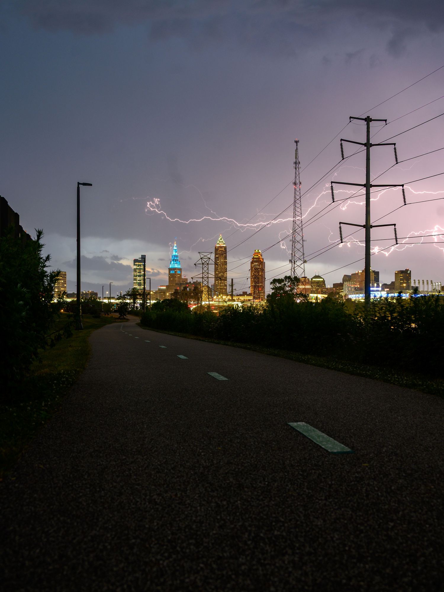 Lightning strikes over Atlanta skyline at dusk with a winding path and power lines in the foreground.