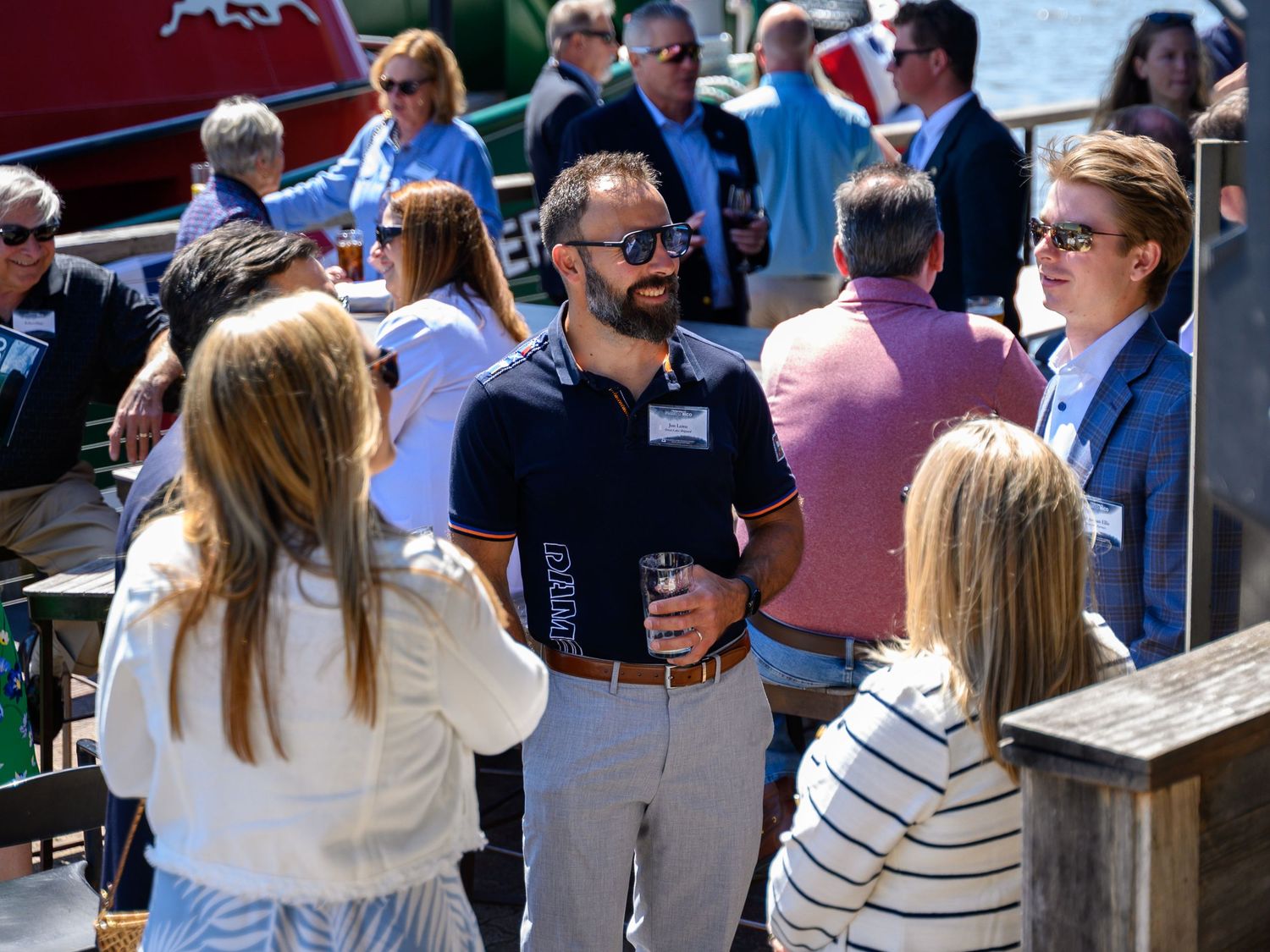Professionals networking outdoors at a waterfront event on a sunny day, socializing with drinks in hand.