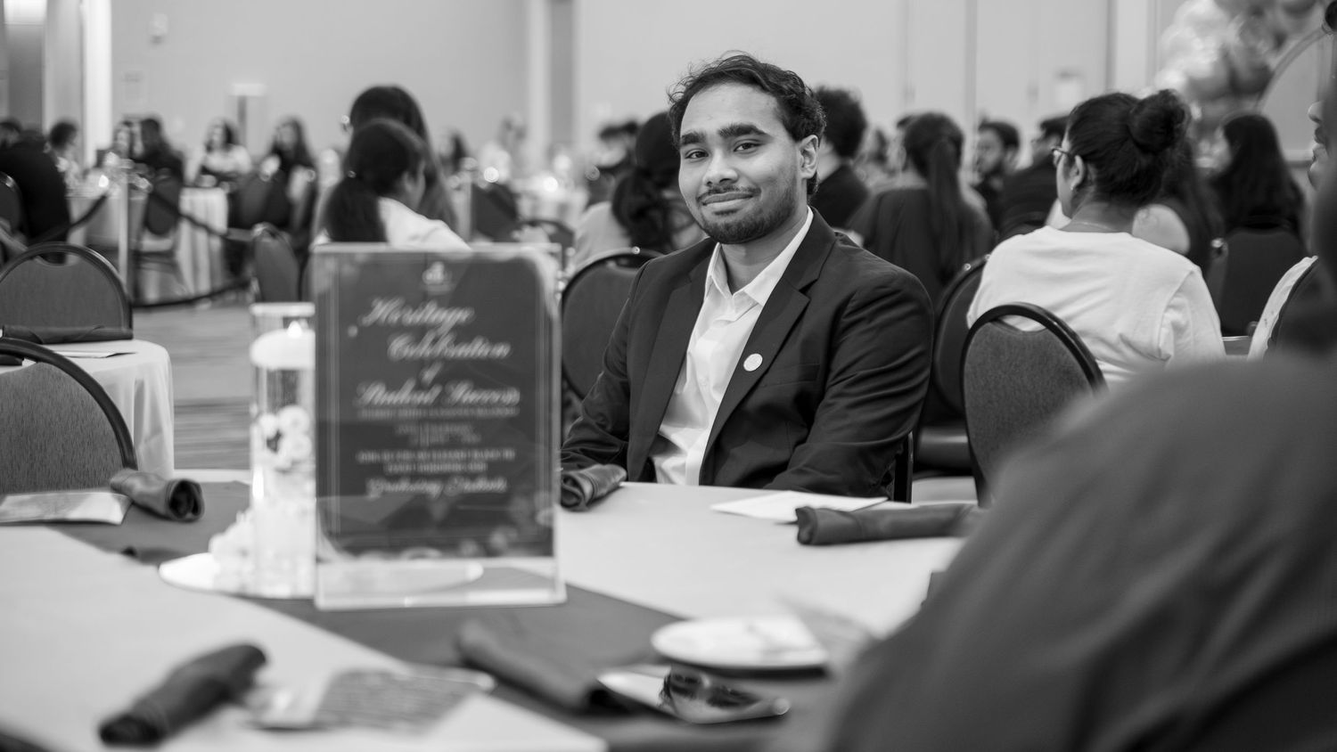 Young man in a suit sitting at a banquet table with an event program card displayed nearby.