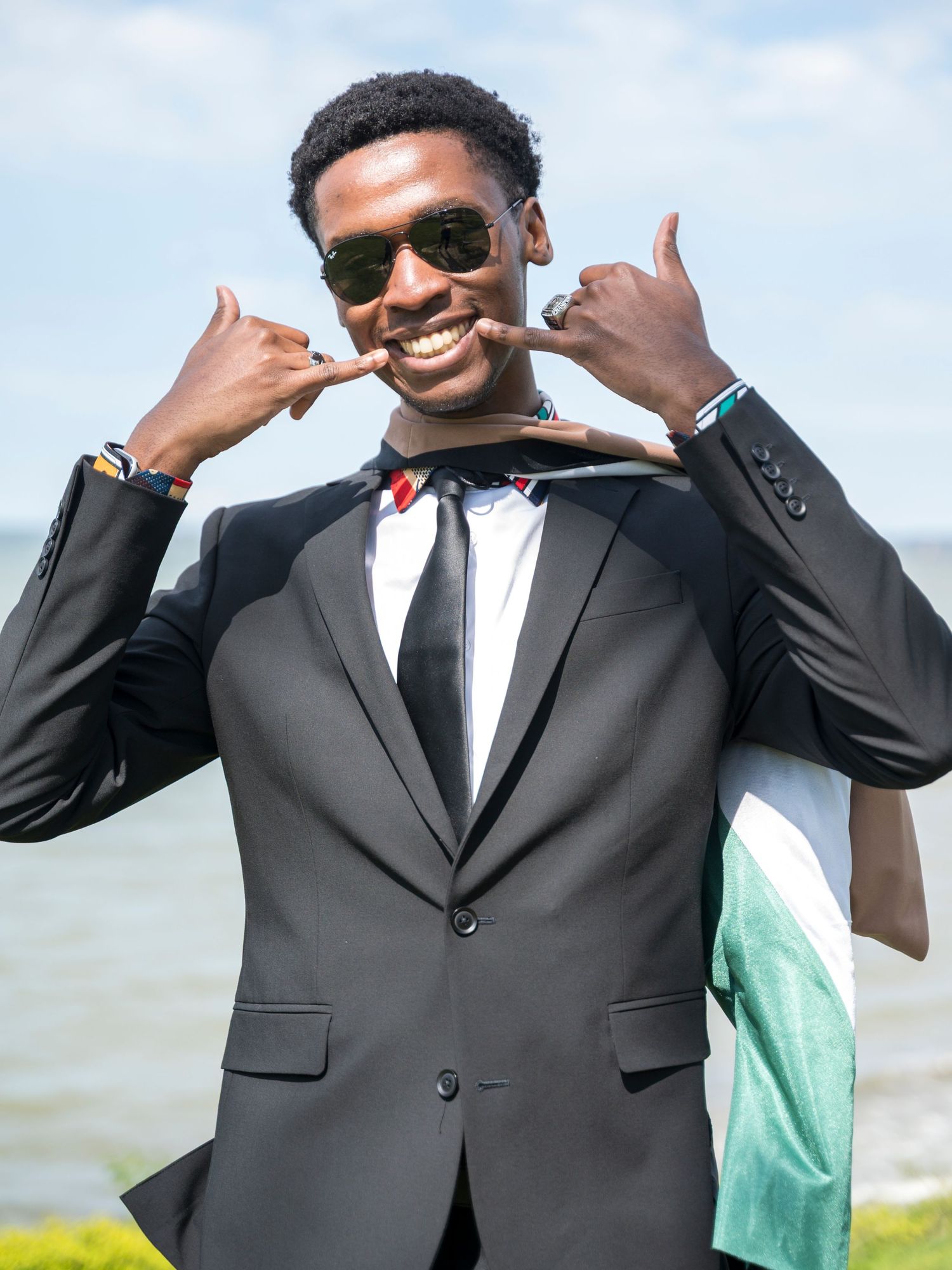 Smiling young man in black suit and bow tie posing outdoors near waterfront on a sunny day.