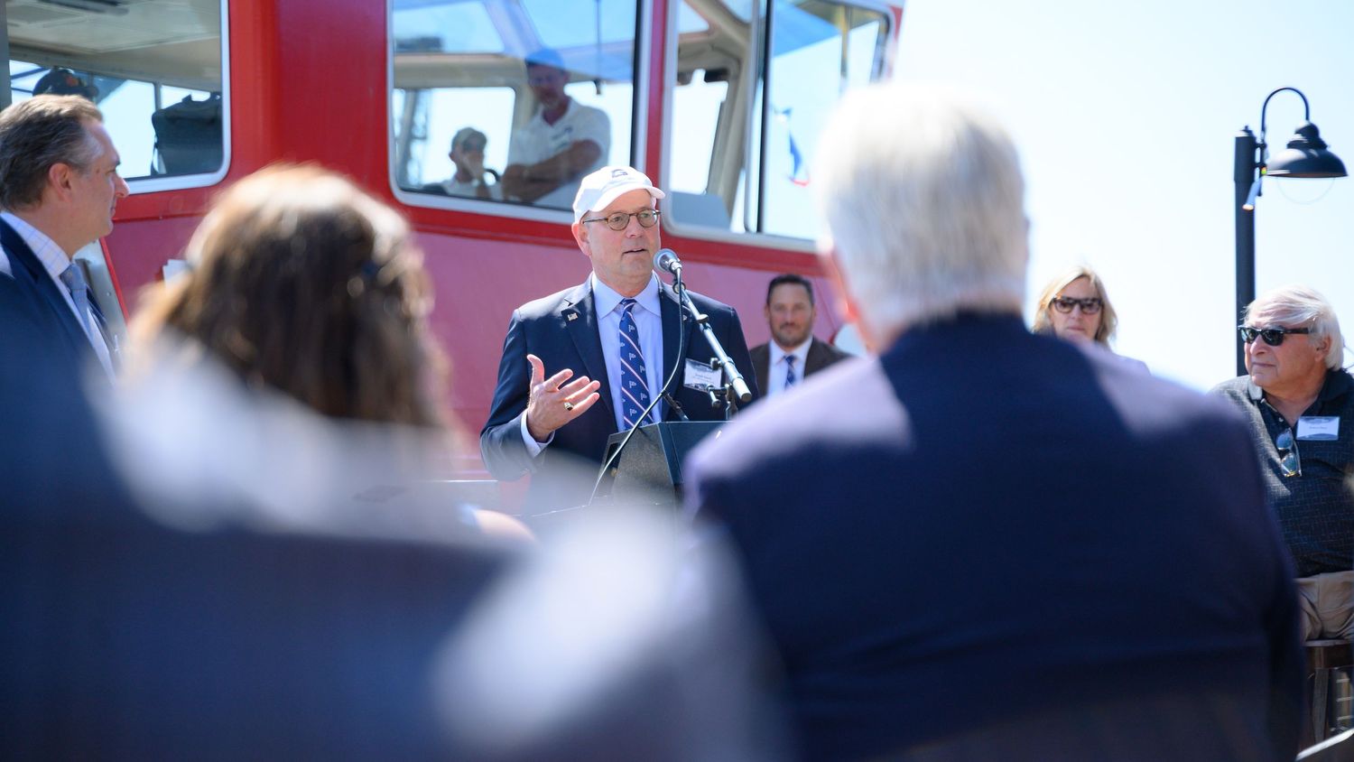 A man in a blue suit speaks at an outdoor event near a red vehicle, surrounded by attentive guests.