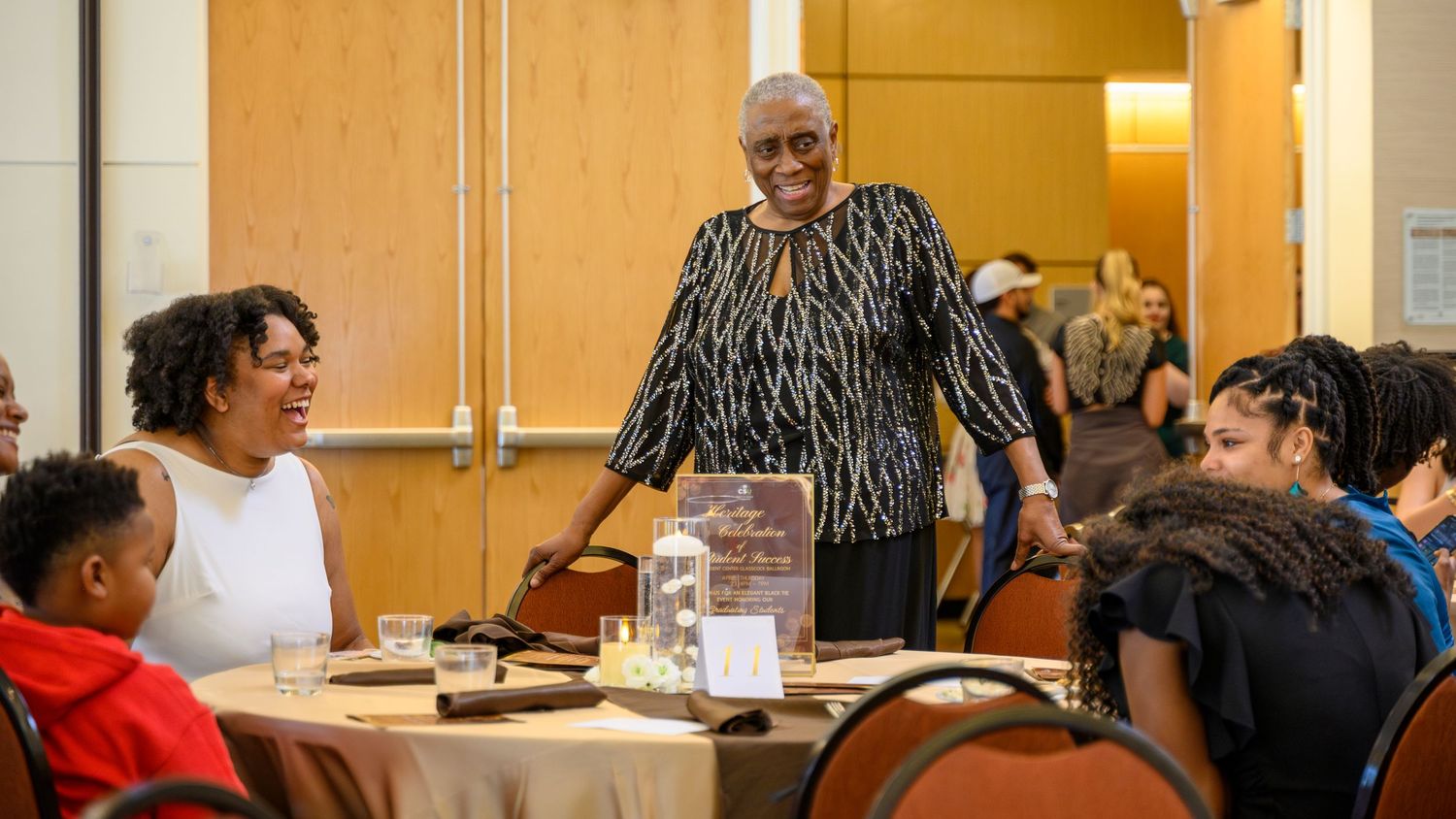 Elderly Black woman standing and smiling at a banquet table with seated guests during a formal gathering.