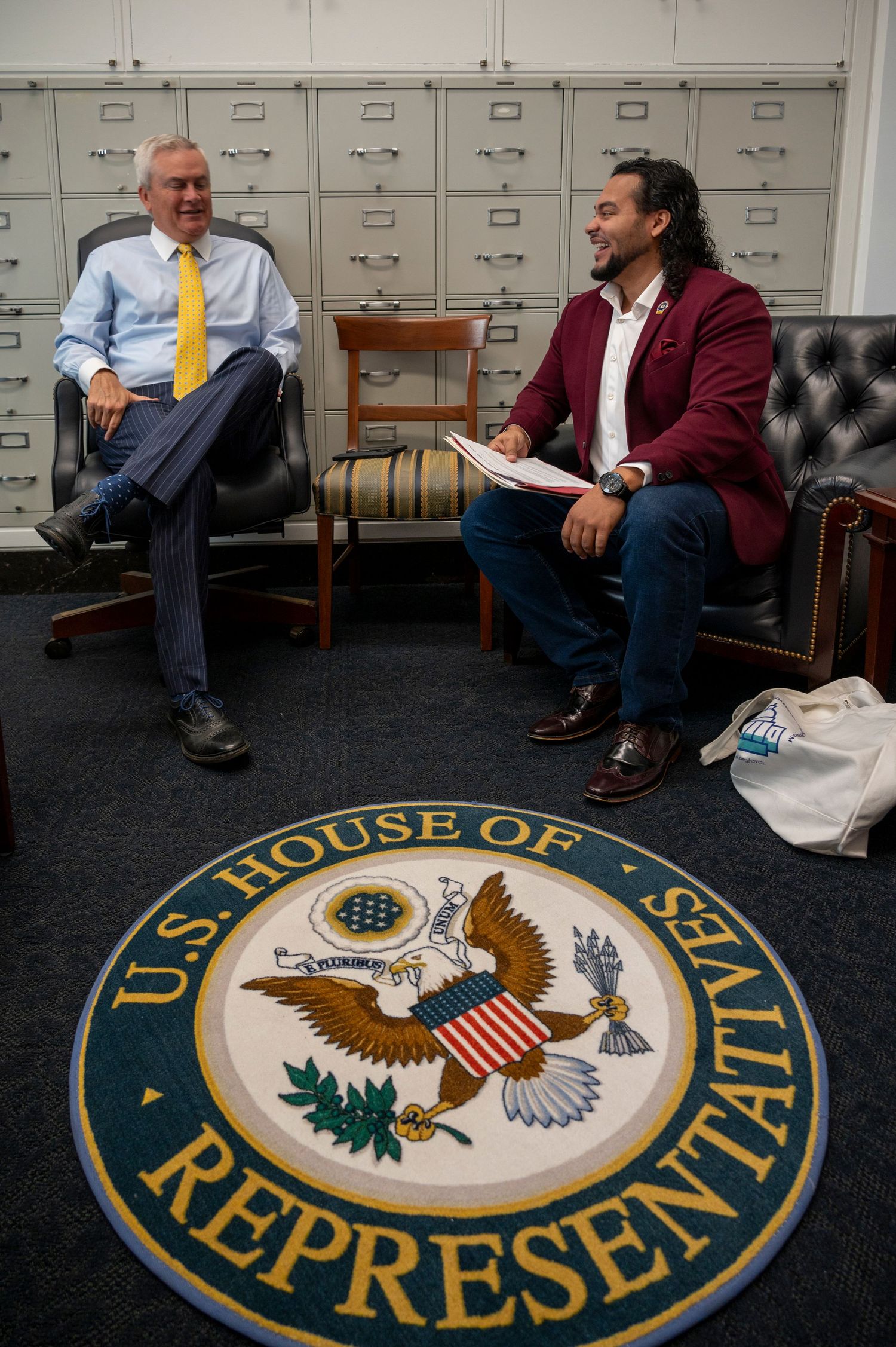 Two men share a laugh in an office with a U.S. House of Representatives seal on the floor.