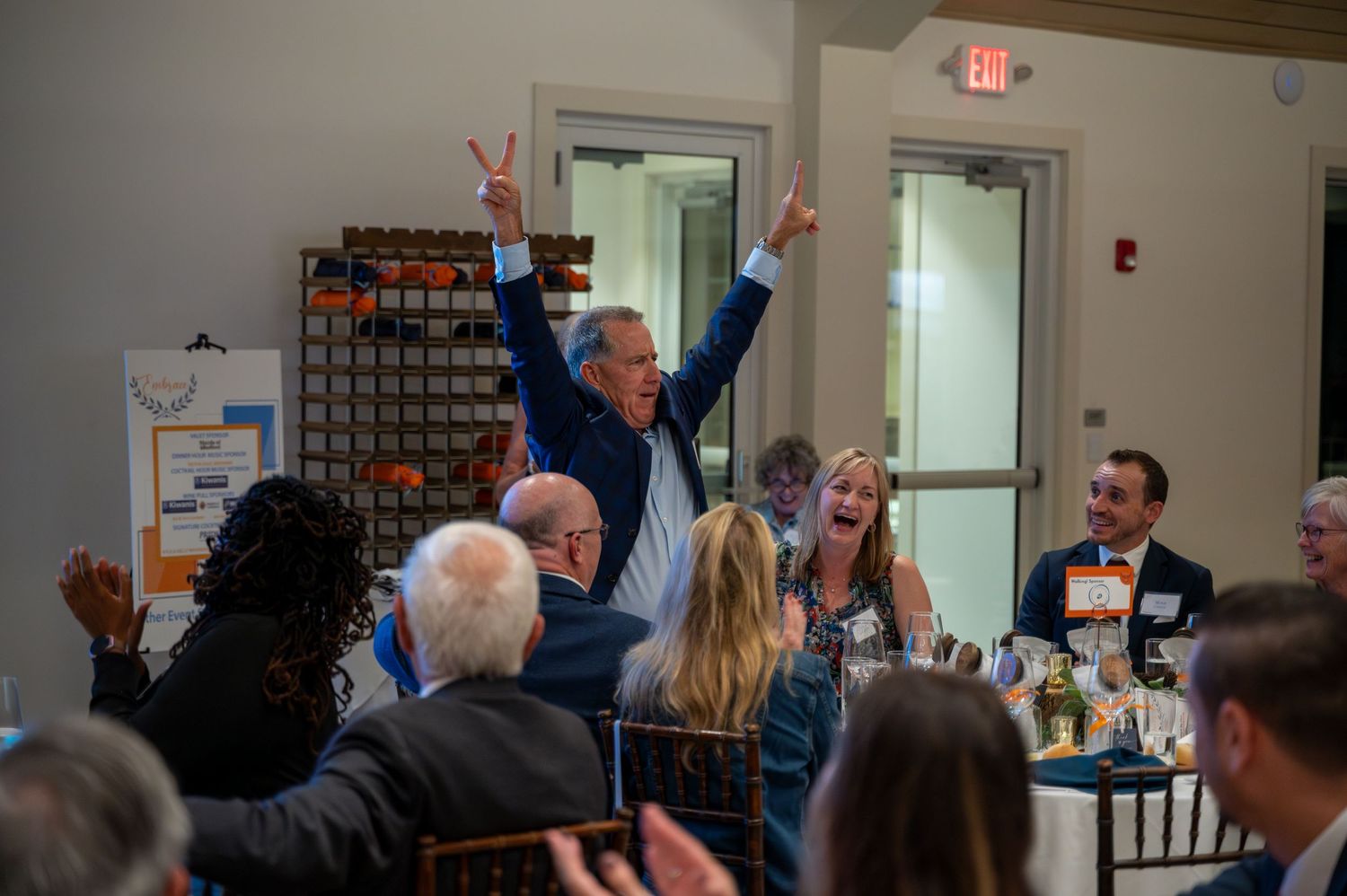 Excited man raises arms in celebration surrounded by laughing guests at formal dinner event.
