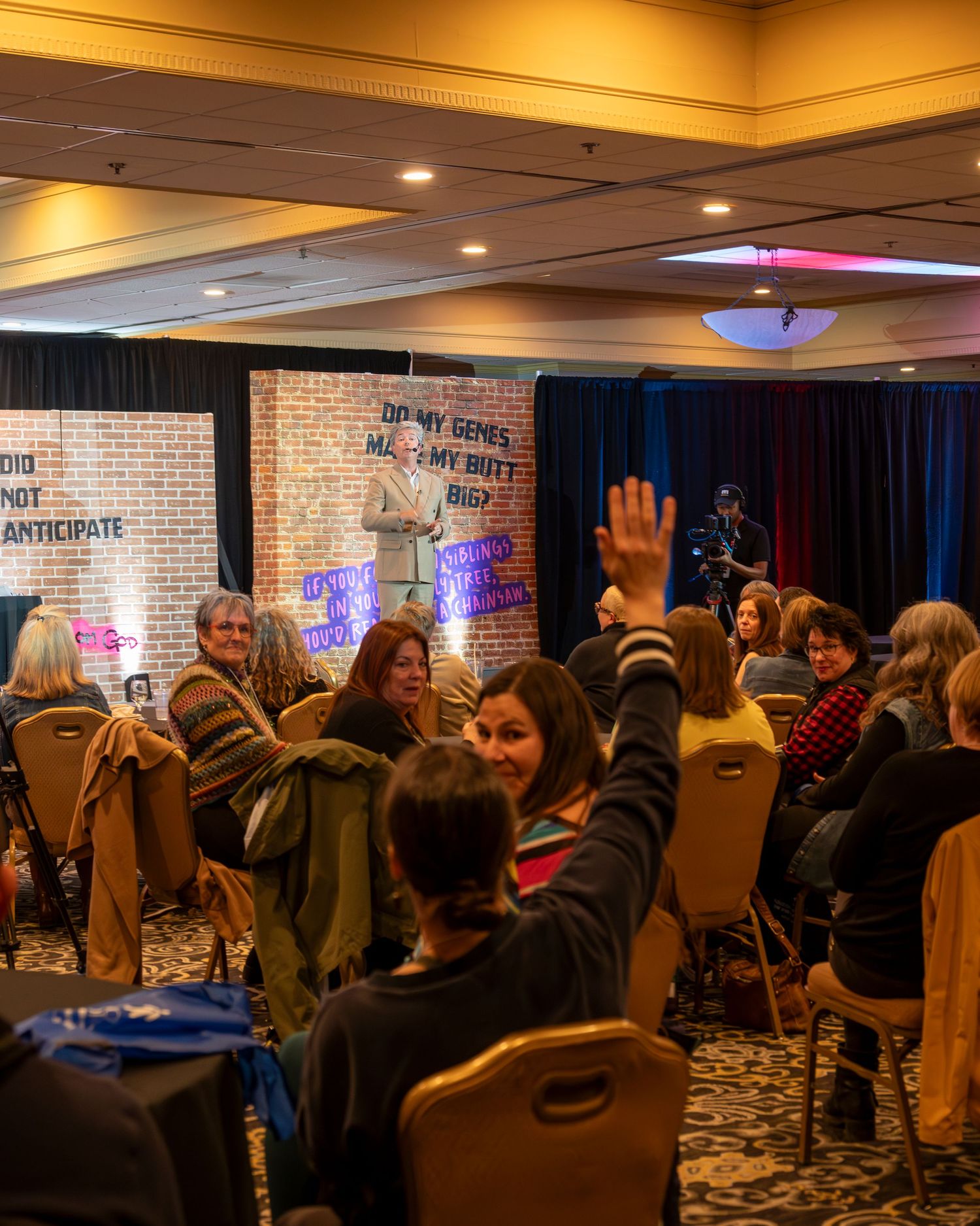Audience members raise hands during a conference session in a hotel ballroom with motivational signage.