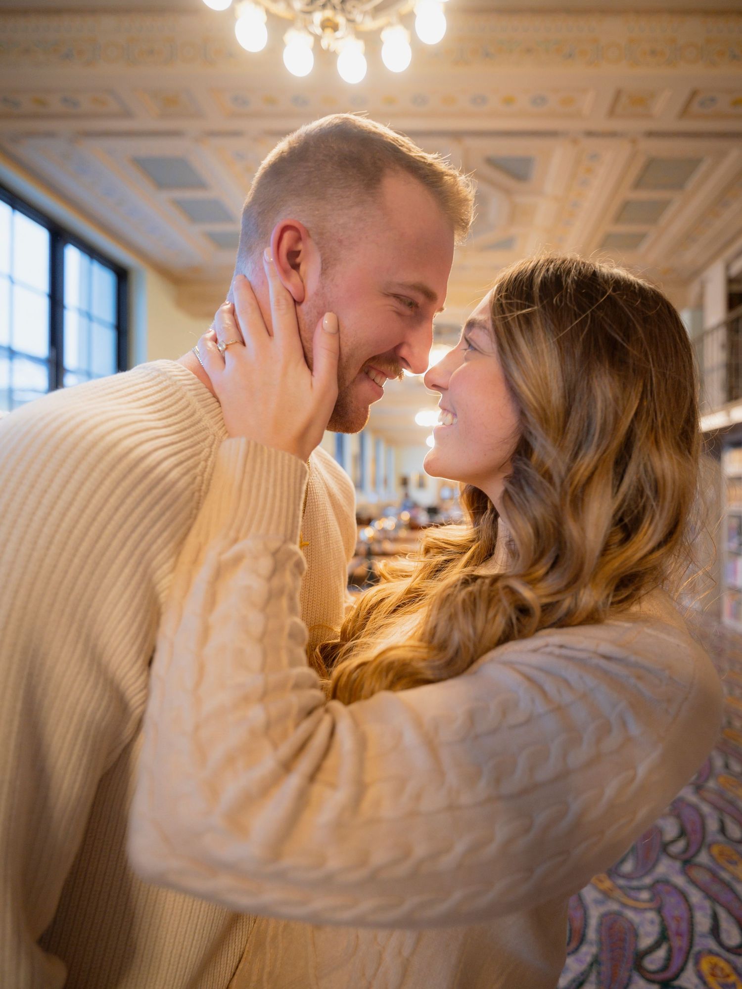 Happy couple in cream sweaters share a romantic moment in an ornate historic building interior.