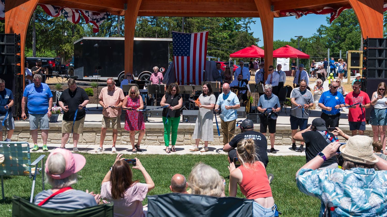 Outdoor community event with crowd seated on lawn watching performers on stage beneath a pavilion with American flag.