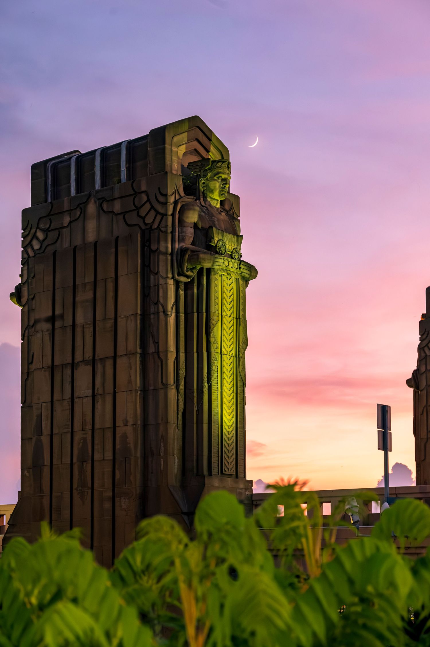 Art Deco stone tower with illuminated guardian statue at sunset, crescent moon visible in vibrant pink and purple sky.