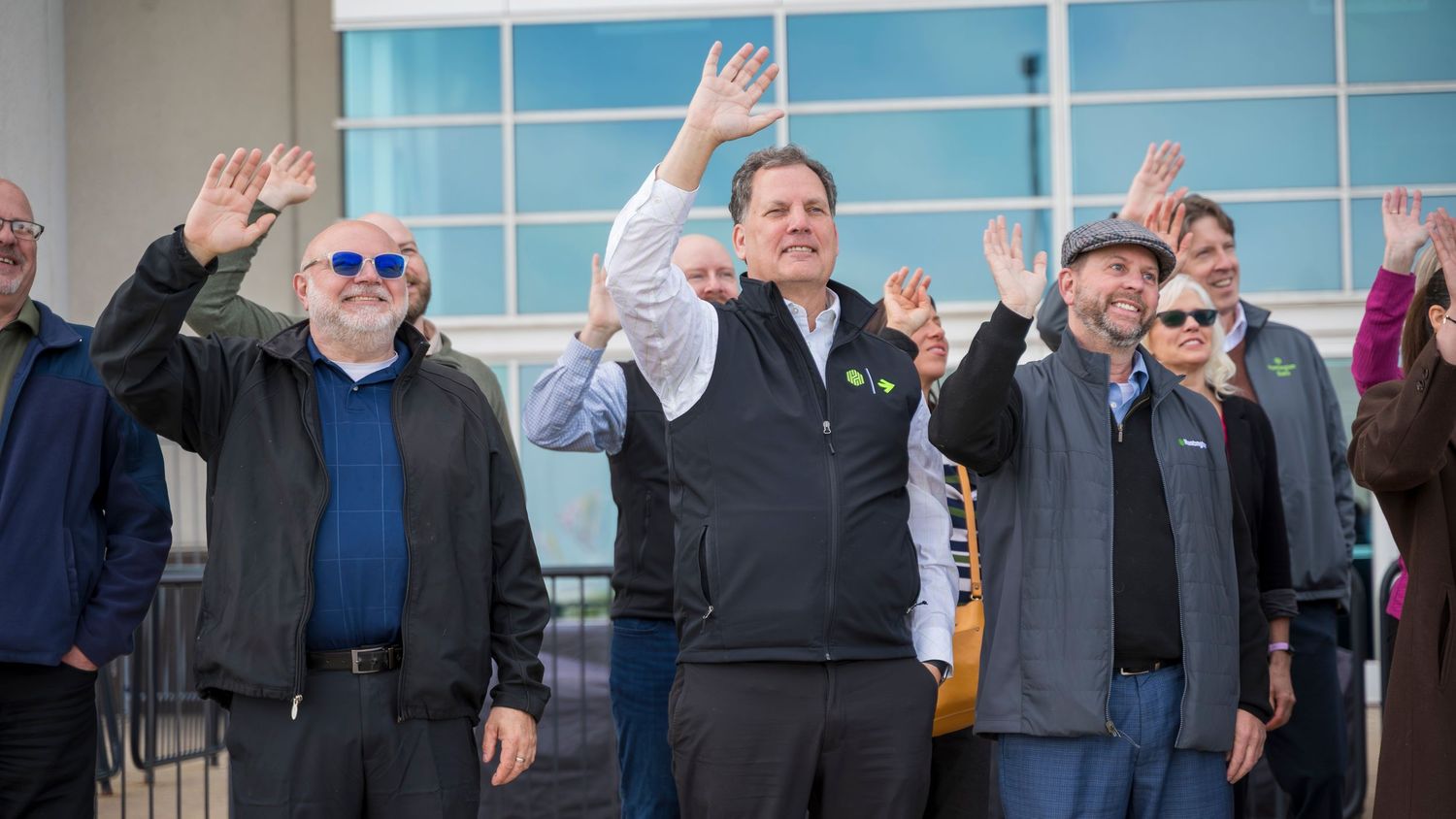 Group of men waving to crowd at outdoor corporate event with smiles and casual attire.
