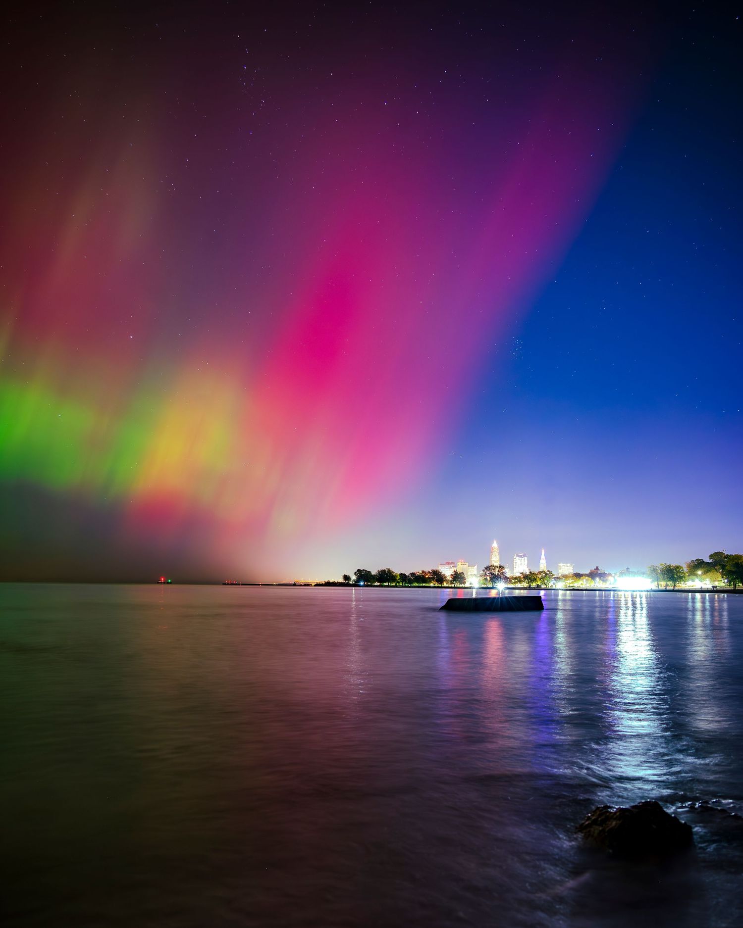 Vibrant aurora borealis with pink, green, and purple lights over a calm lake near a city skyline at night.