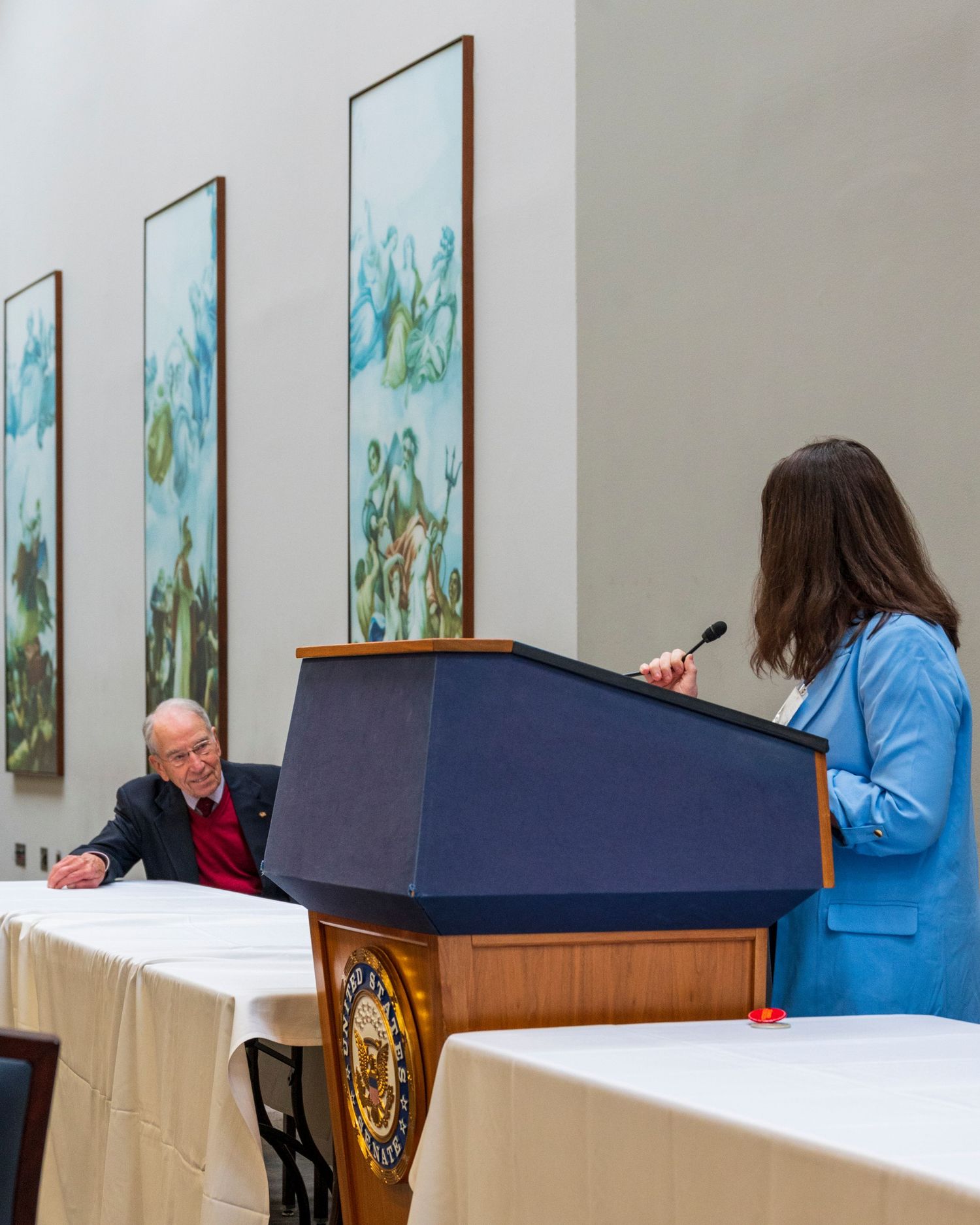 A woman in a blue blazer speaks at a navy podium before an elderly seated listener in a formal meeting room.