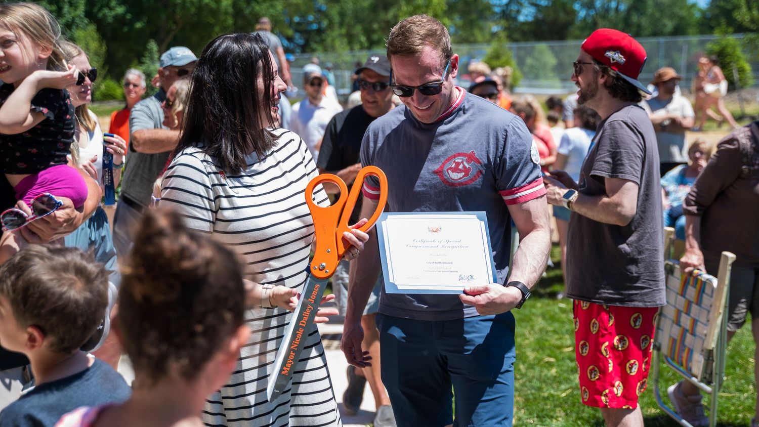Man holding certificate and orange scissors at outdoor ribbon-cutting ceremony surrounded by crowd on sunny day.