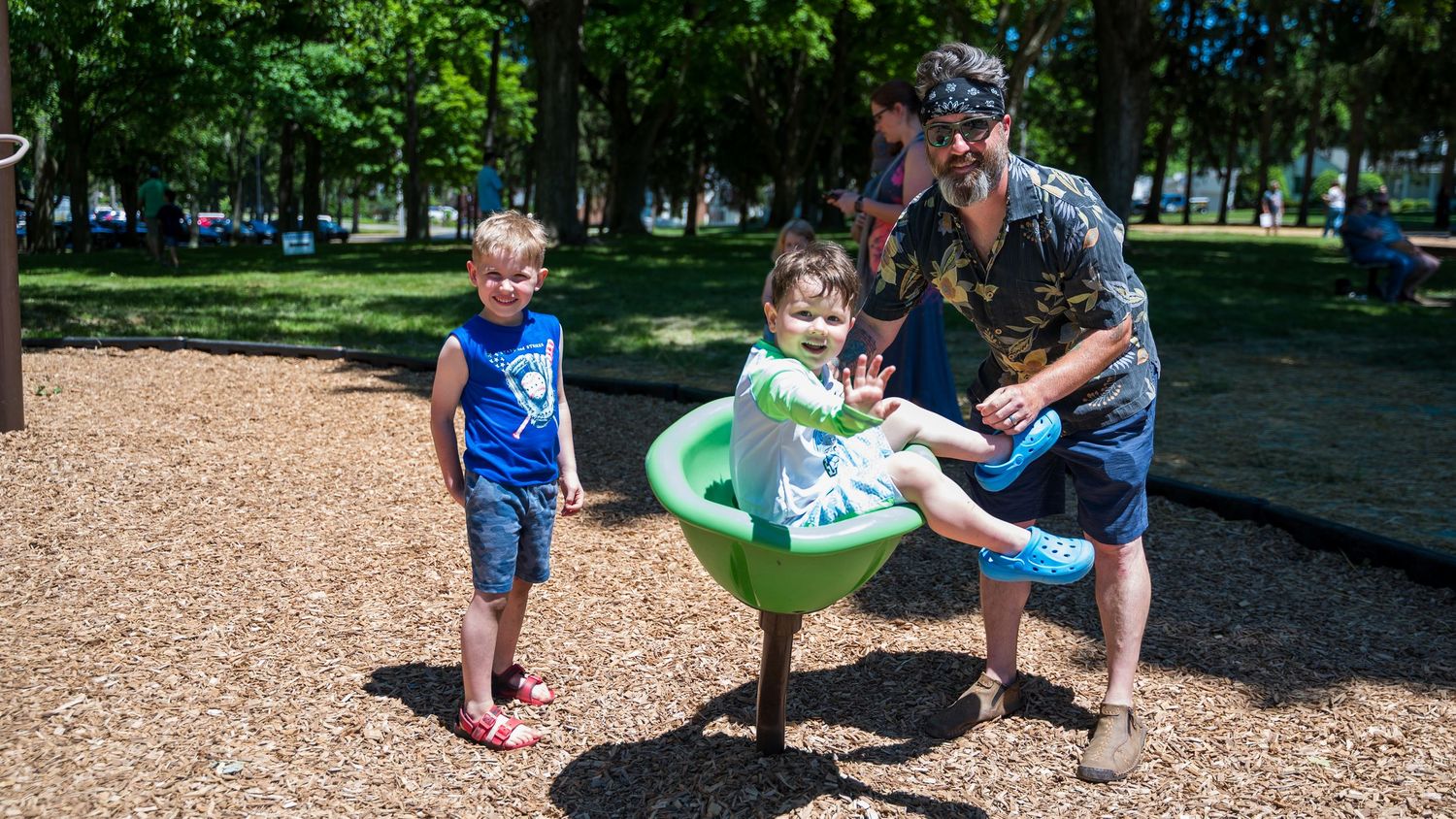 A father helps two young children play on a spinning bowl ride at a sunny outdoor playground surrounded by trees.