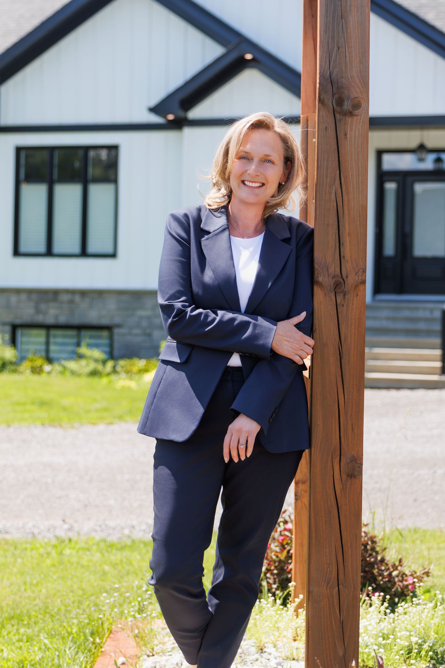 Professional female real estate agent in navy blazer leaning against a wooden post outside a house.
