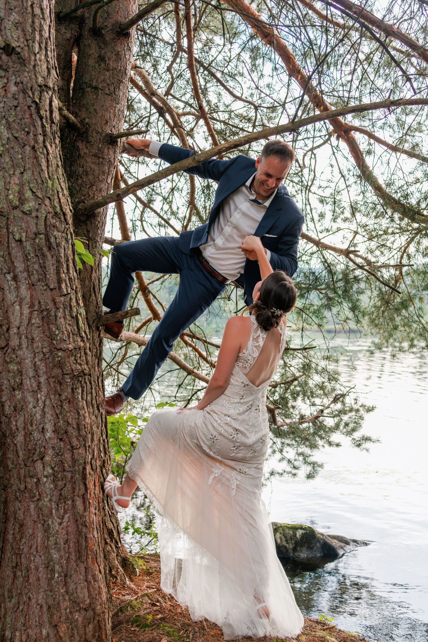 Playful wedding photo of groom climbing tree while bride assists near a lakeside setting.