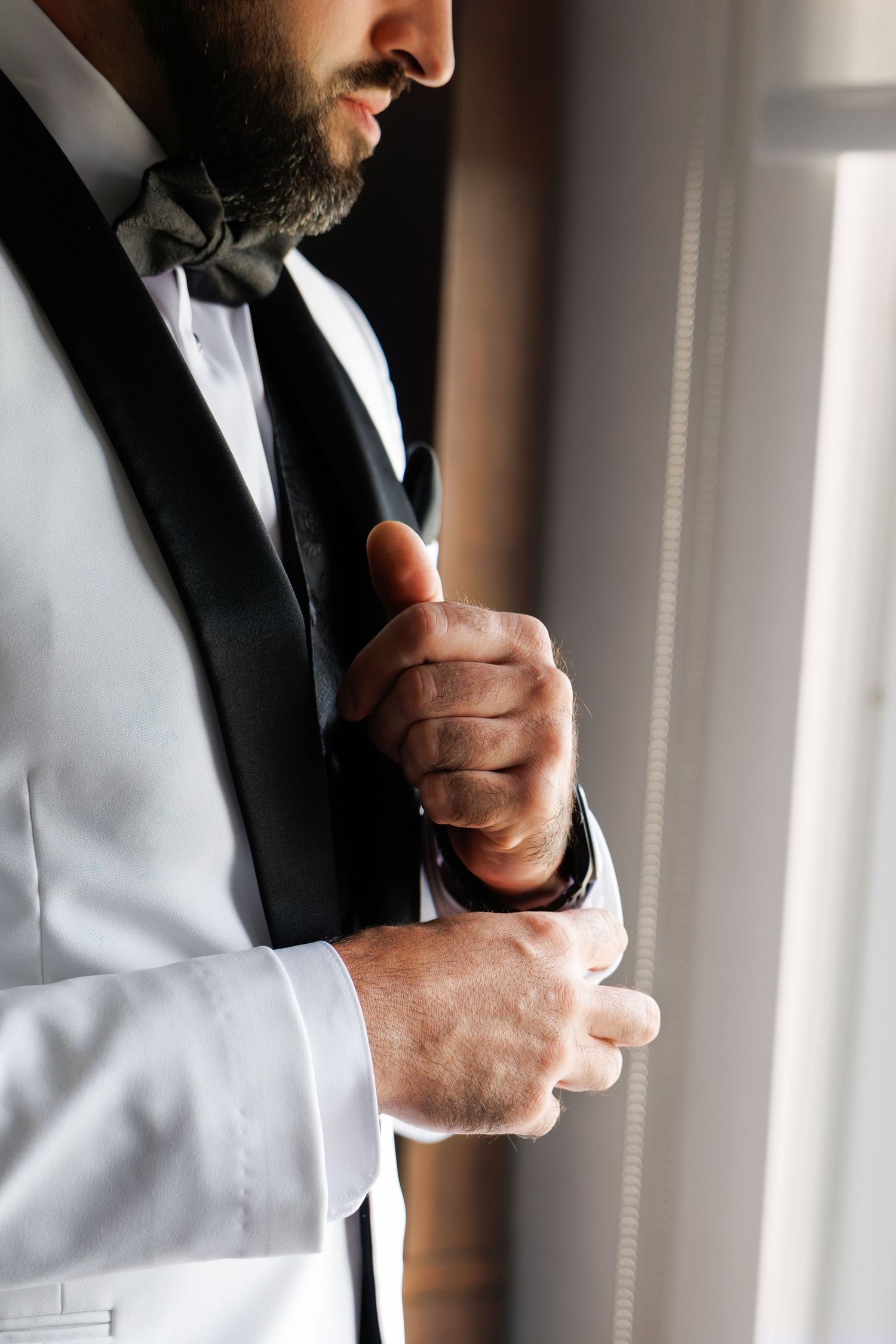 Close-up of groom adjusting his black tuxedo vest and bow tie near a window.