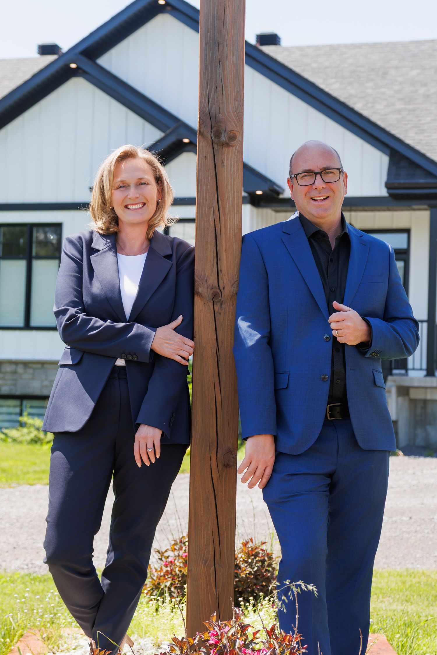 Two professional real estate agents in business suits smiling beside a wooden post outside a home.