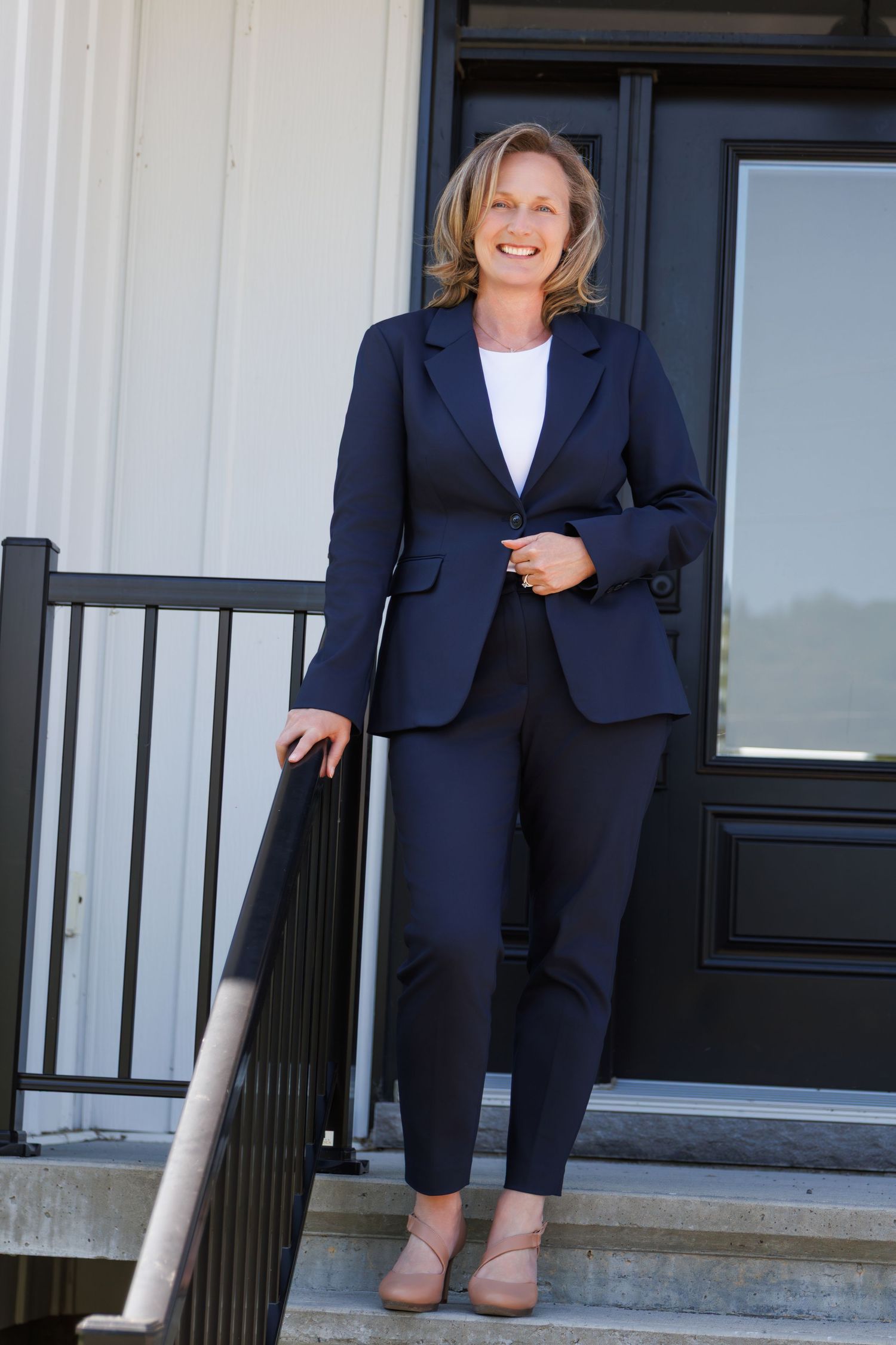 Smiling professional woman in navy business suit standing on front porch steps of a white building.
