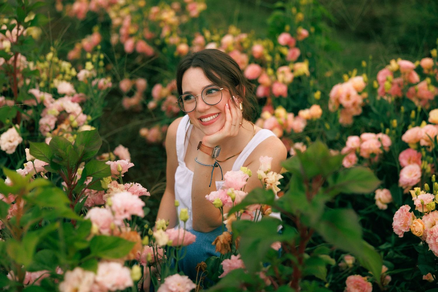Woman with glasses smiling among pink roses in a lush garden, wearing a white top and jeans.
