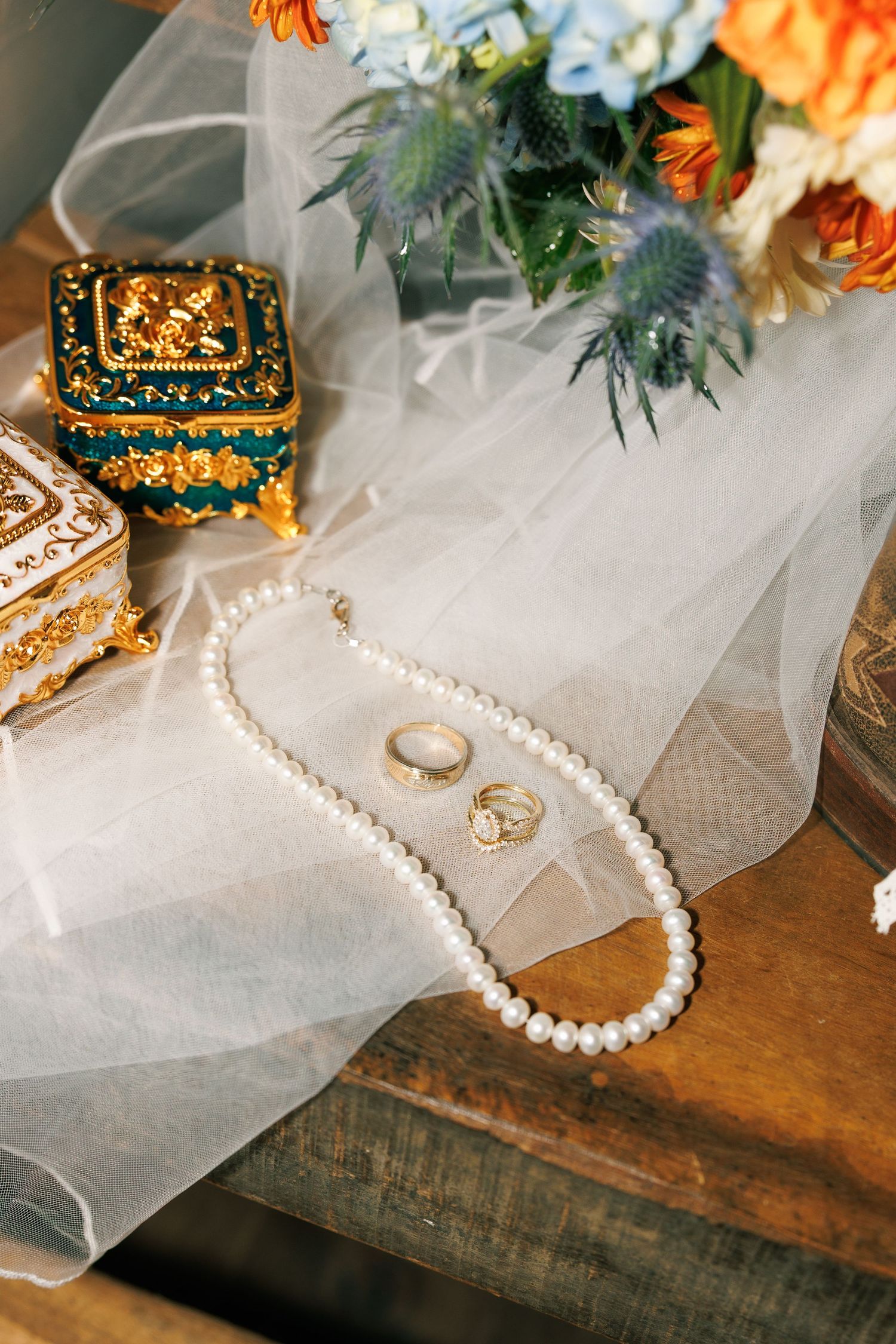 Wedding accessories including pearl necklace and veil arranged on rustic wooden surface with orange flowers.