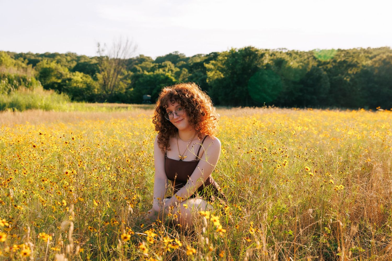A young woman with curly red hair sits among yellow wildflowers in a sunlit meadow wearing a brown sundress.