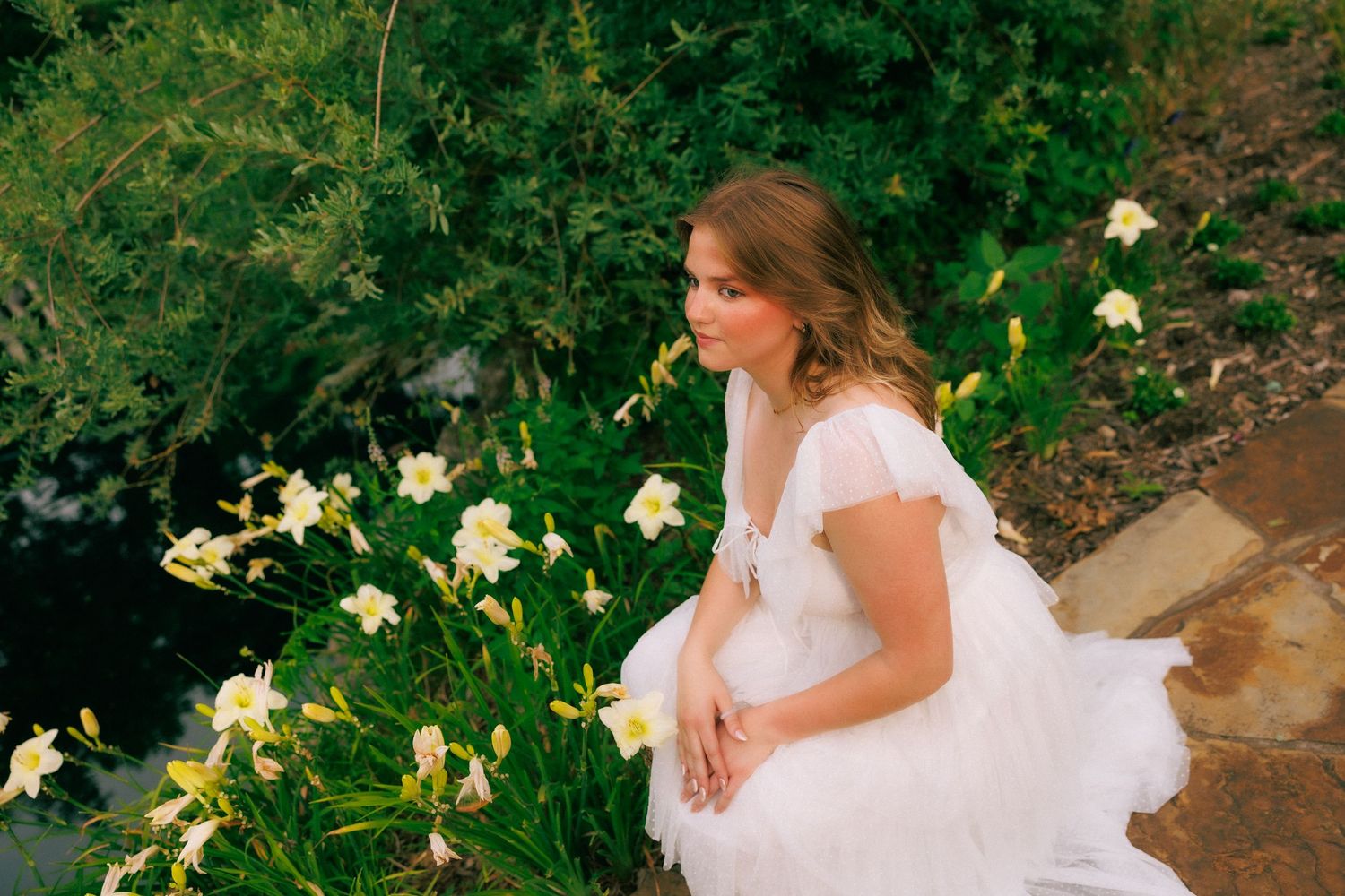 Bride in white gown sitting among yellow daylilies in lush green garden, elegant outdoor wedding photography.