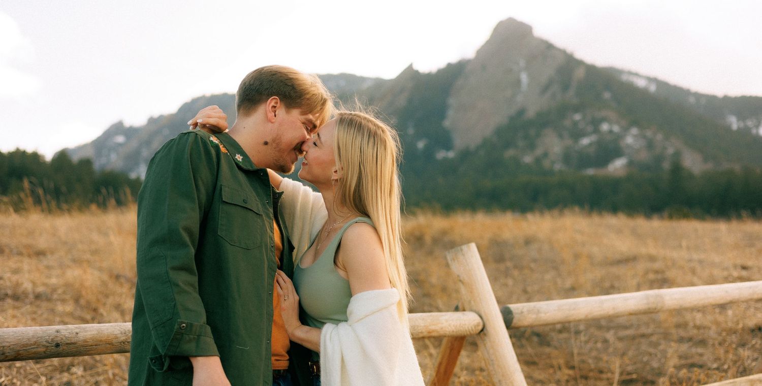 Couple sharing a romantic moment near a wooden fence with scenic mountain backdrop in an open field.