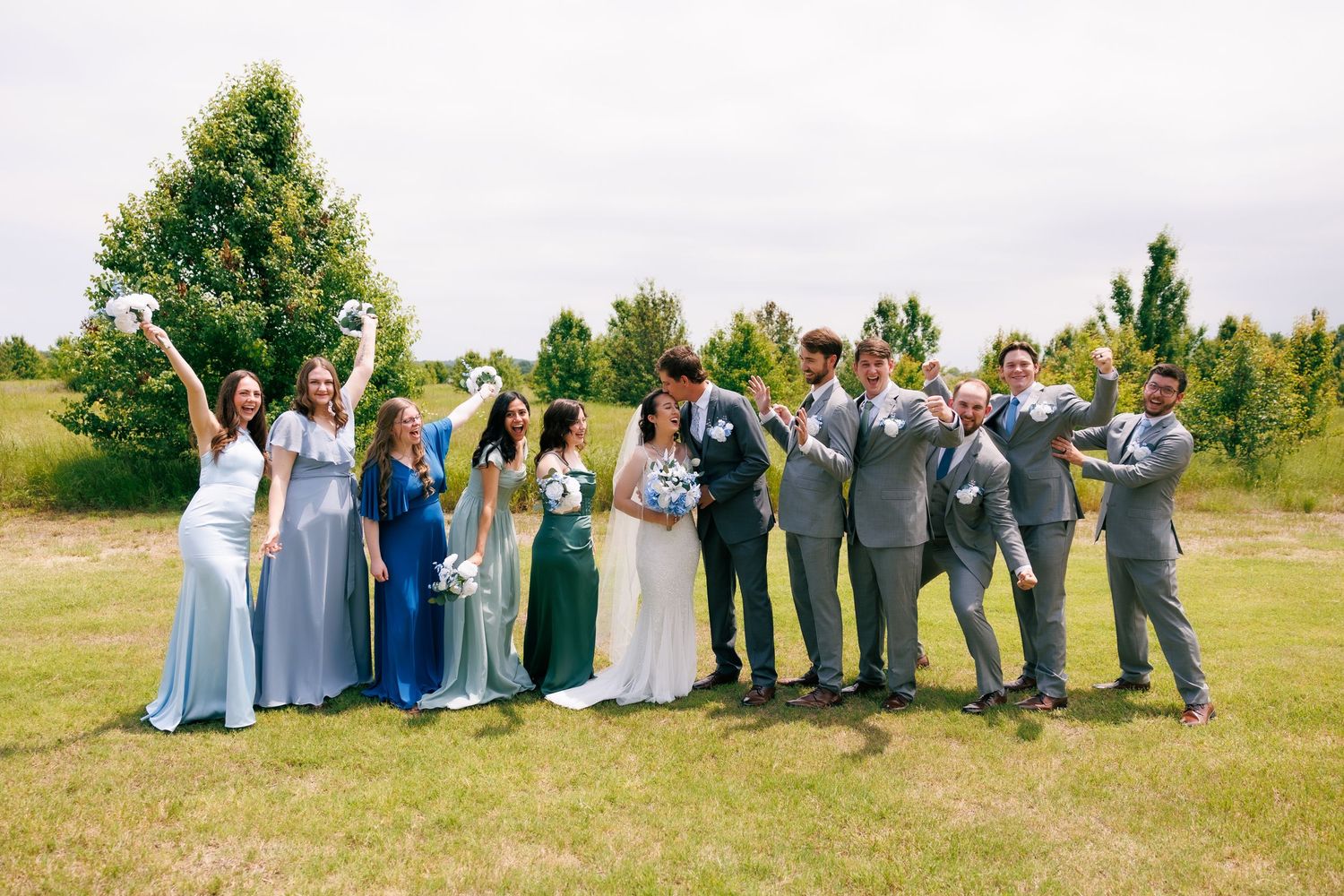 Large wedding party poses together outdoors wearing coordinated blue and grey attire.