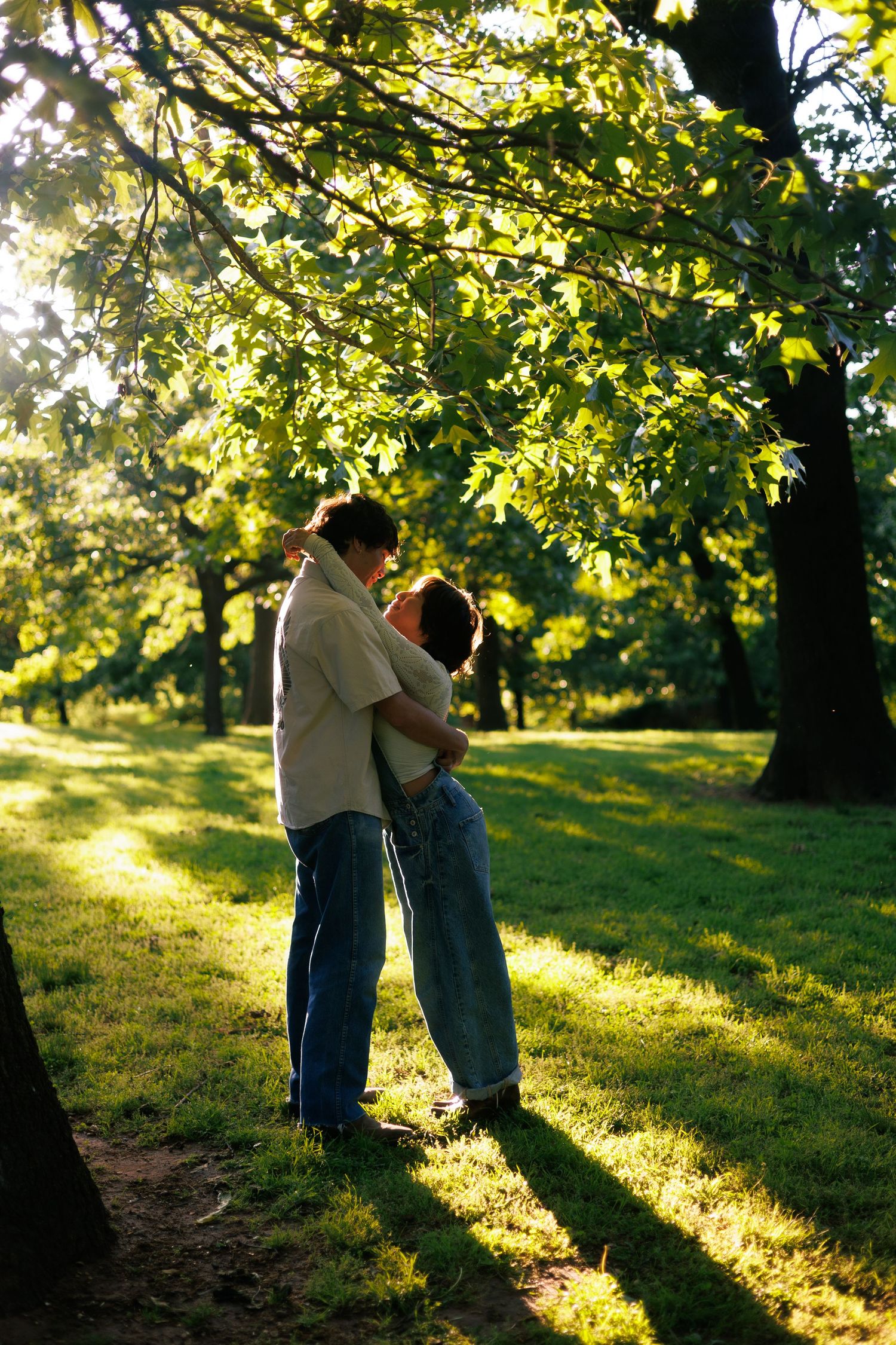 A romantic moment captured in a sunlit park with trees casting long shadows.