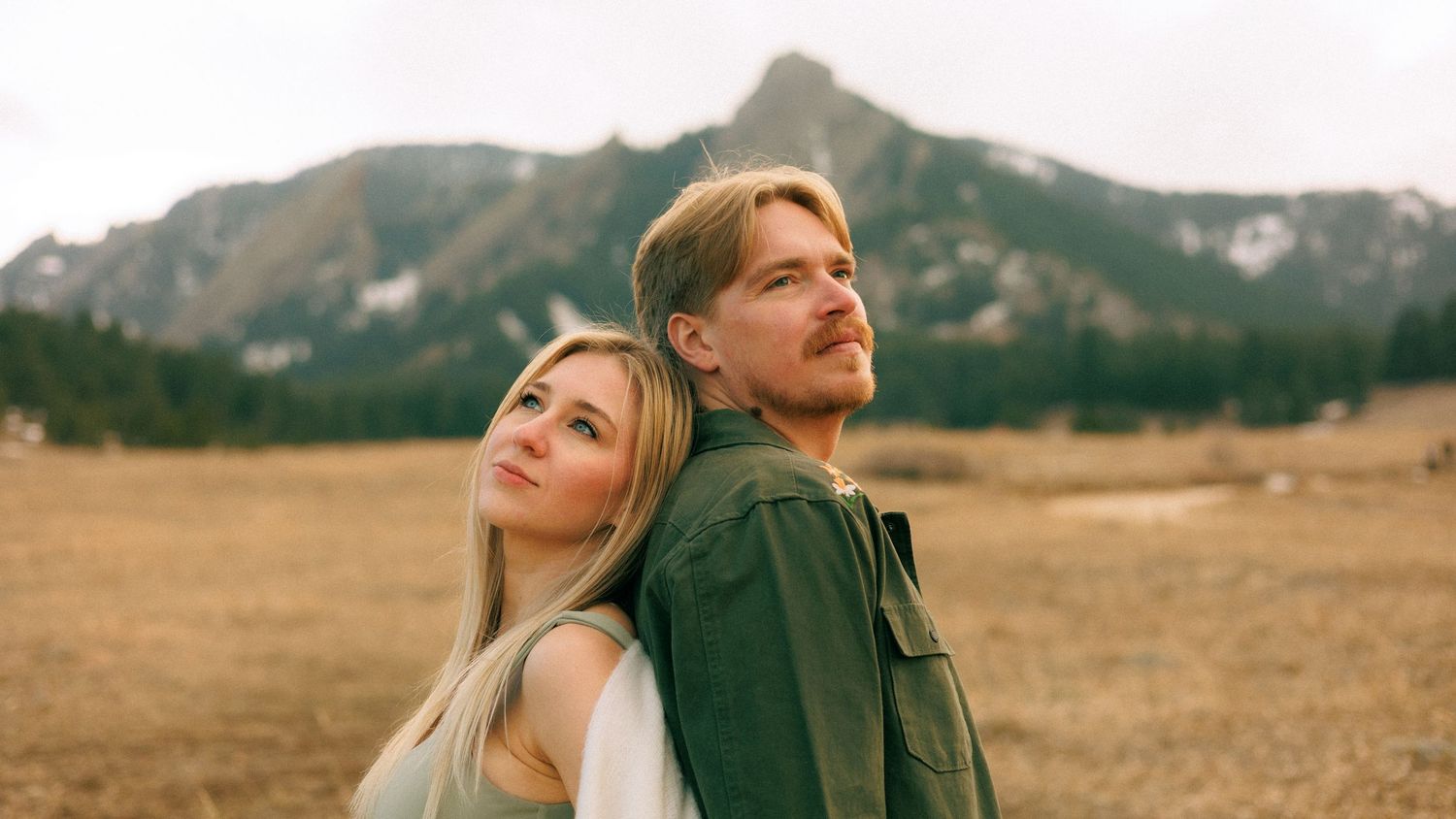A couple stands back-to-back in a mountain meadow, the woman with blonde hair and man in a green jacket gazing away.