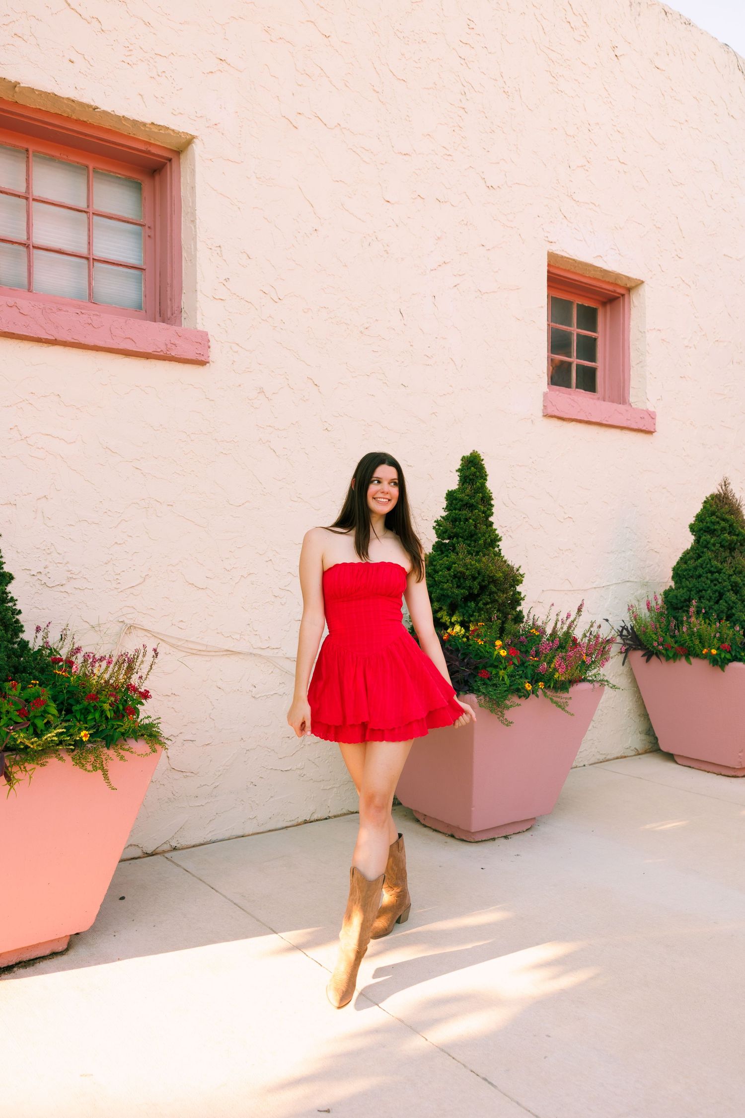 A red sundress and brown boots pop against a pink stucco wall with window details.
