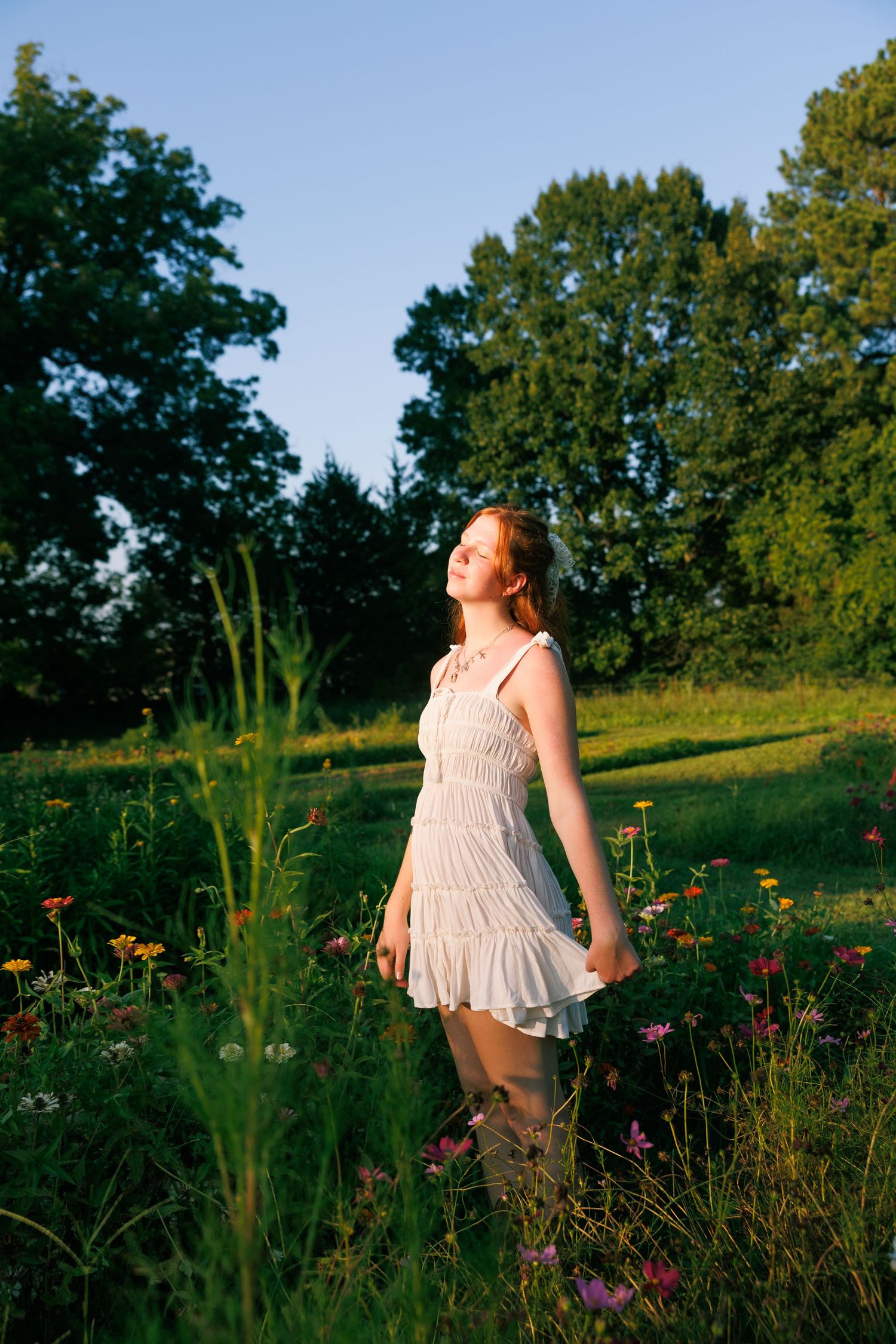 A white sundress-clad figure stands in a wildflower meadow at golden hour with trees in the background.