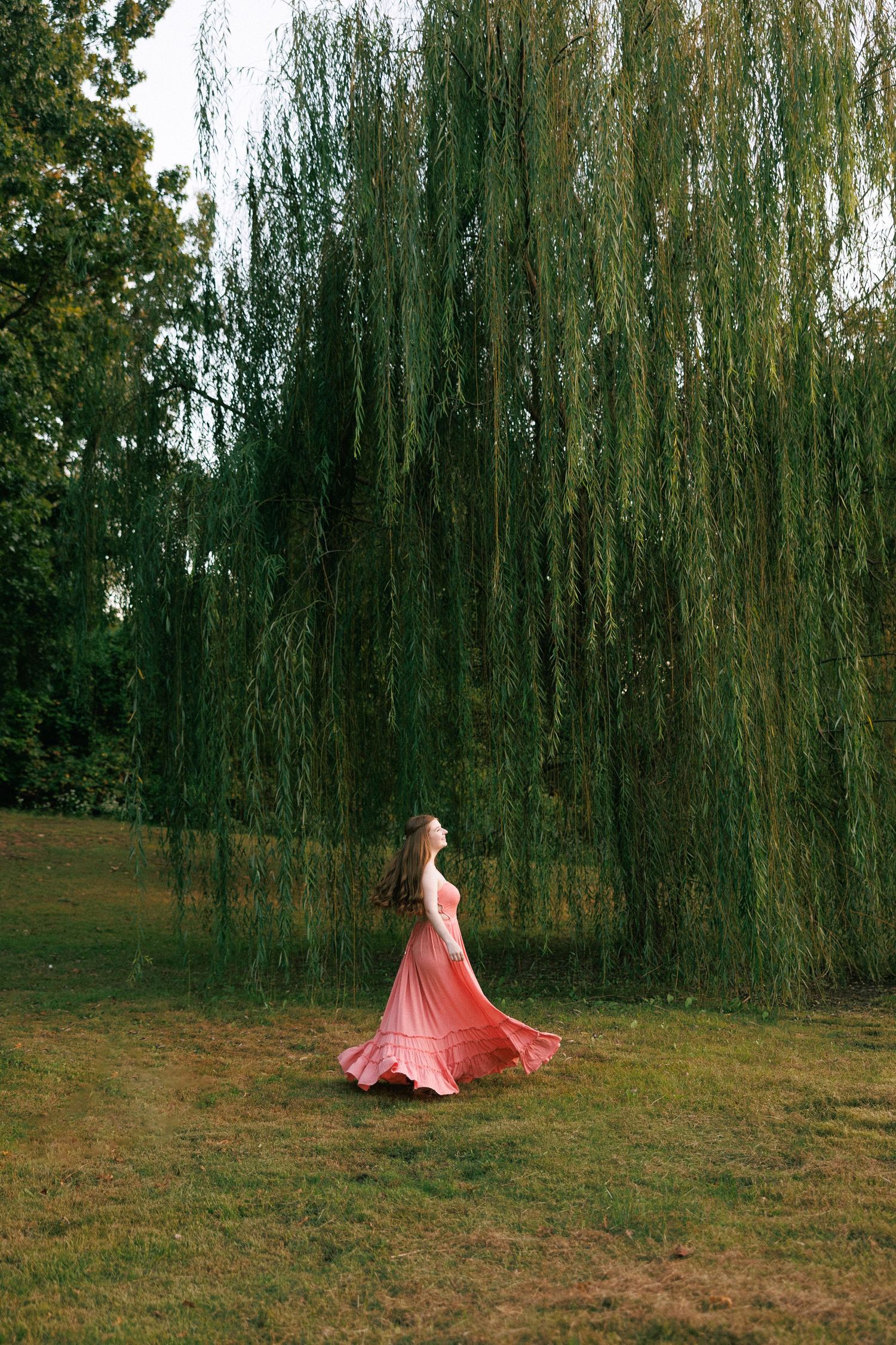 A flowing coral dress twirls against a backdrop of weeping willow trees in a park setting.