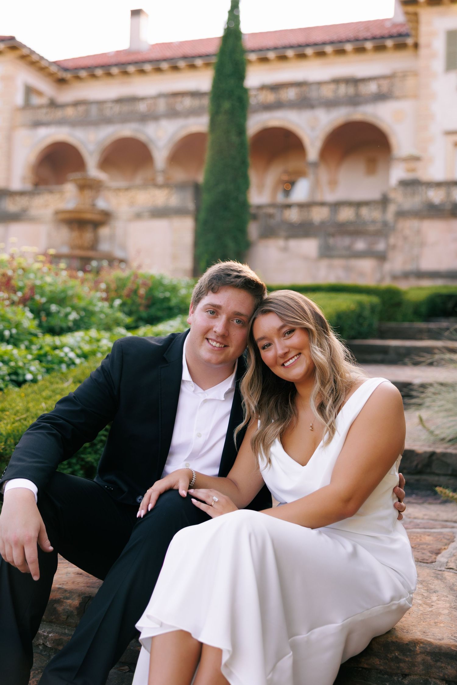 A couple in formal attire shares intimate moments on stone steps with cypress trees in the background.