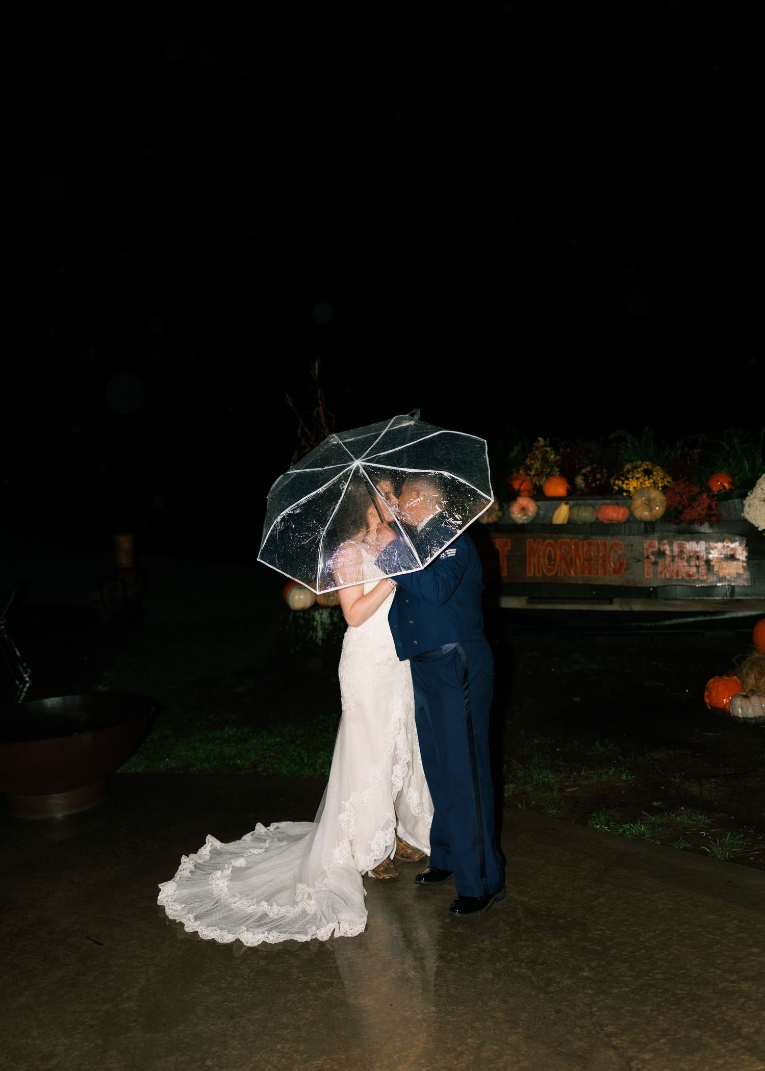 Couple shares romantic moment under clear umbrella at night with orange lights in background.