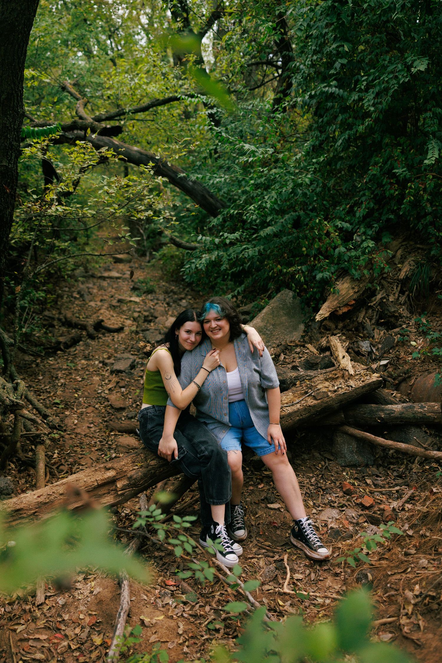 Two people sitting close together on fallen leaves in a wooded forest setting.