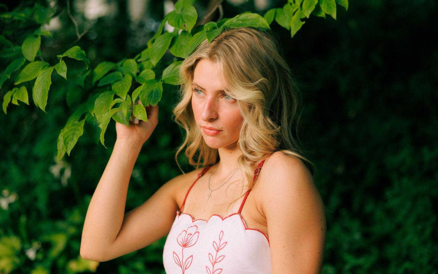 Young blonde woman in white floral top poses gracefully among lush green tree branches outdoors.