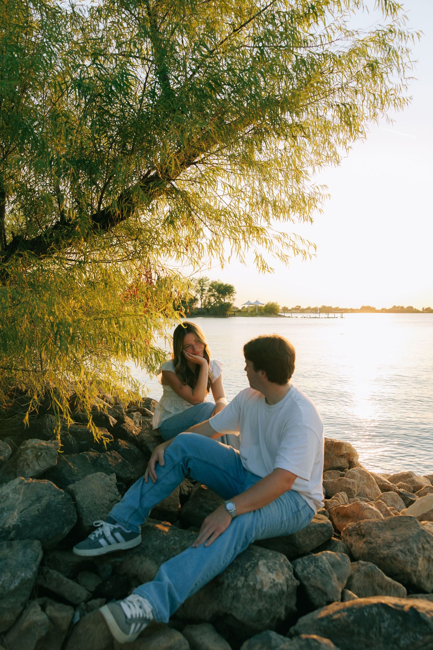 Couple sitting on rocks by a sunlit river under a willow tree, enjoying a golden hour moment together.