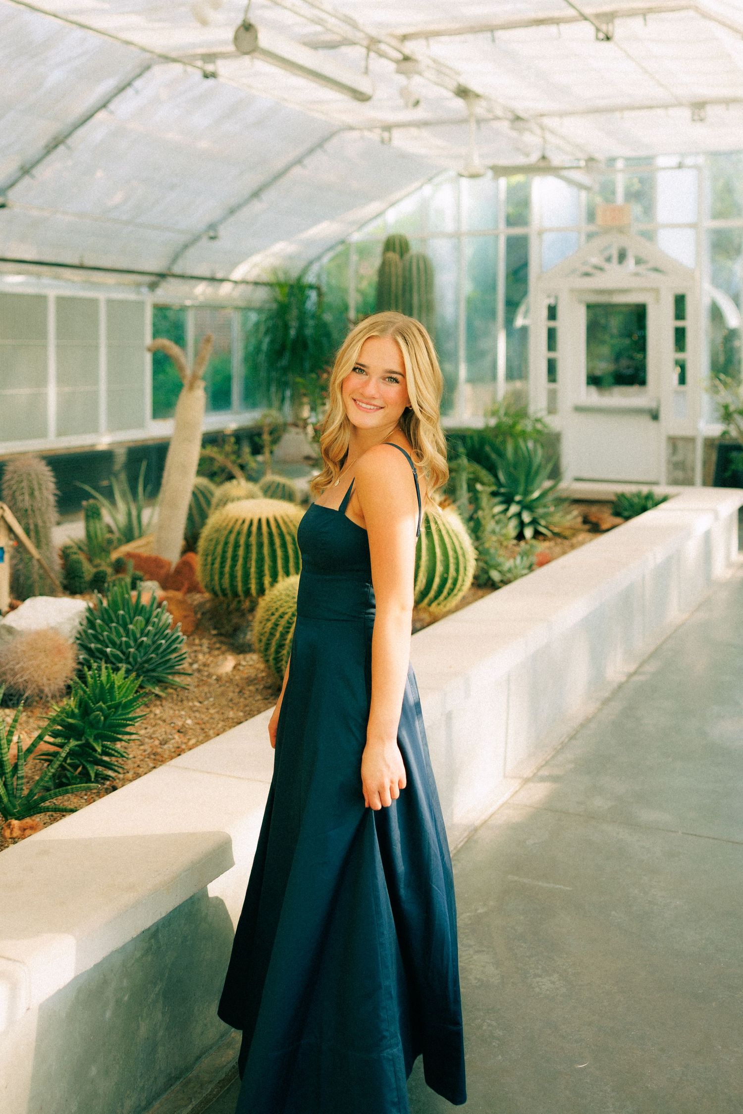 Young woman in elegant navy gown posing in a greenhouse with cacti and succulents in the background.