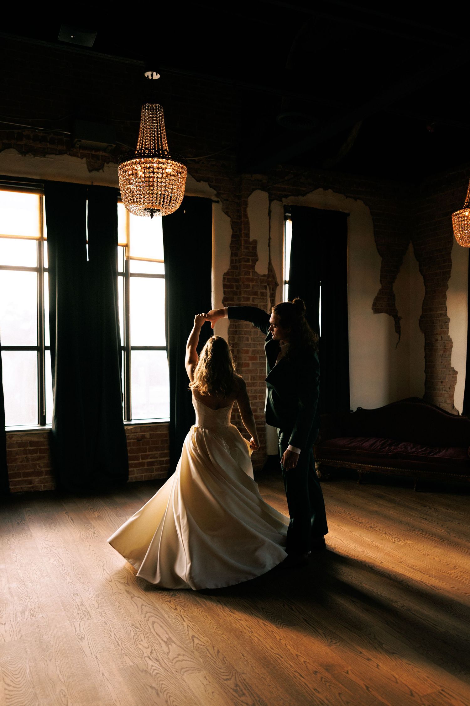 A couple dances in silhouette against dramatic window light in an elegant room with chandeliers.