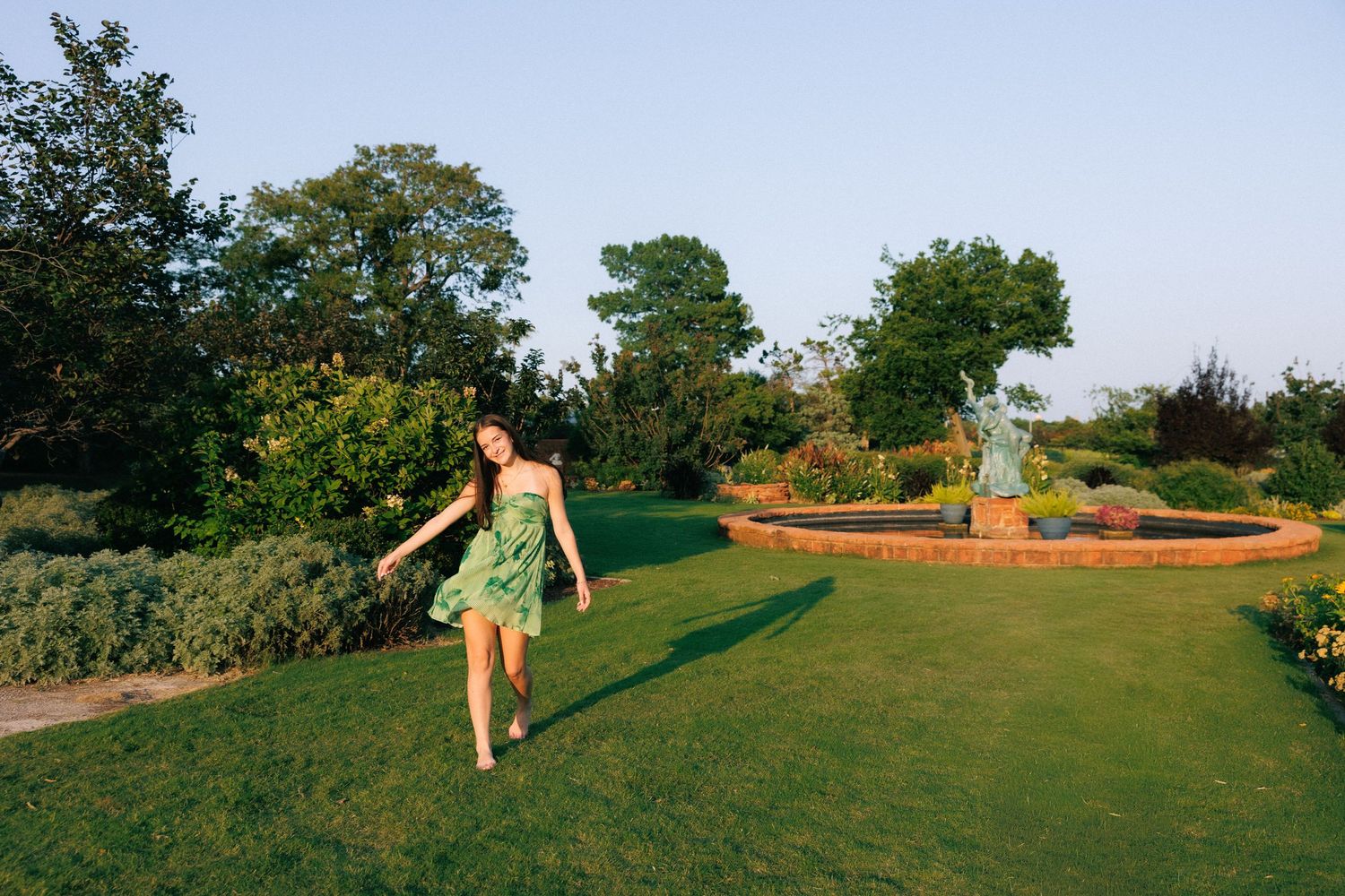 Woman in green dress walking on lush garden lawn with fountain and trees in golden hour light.