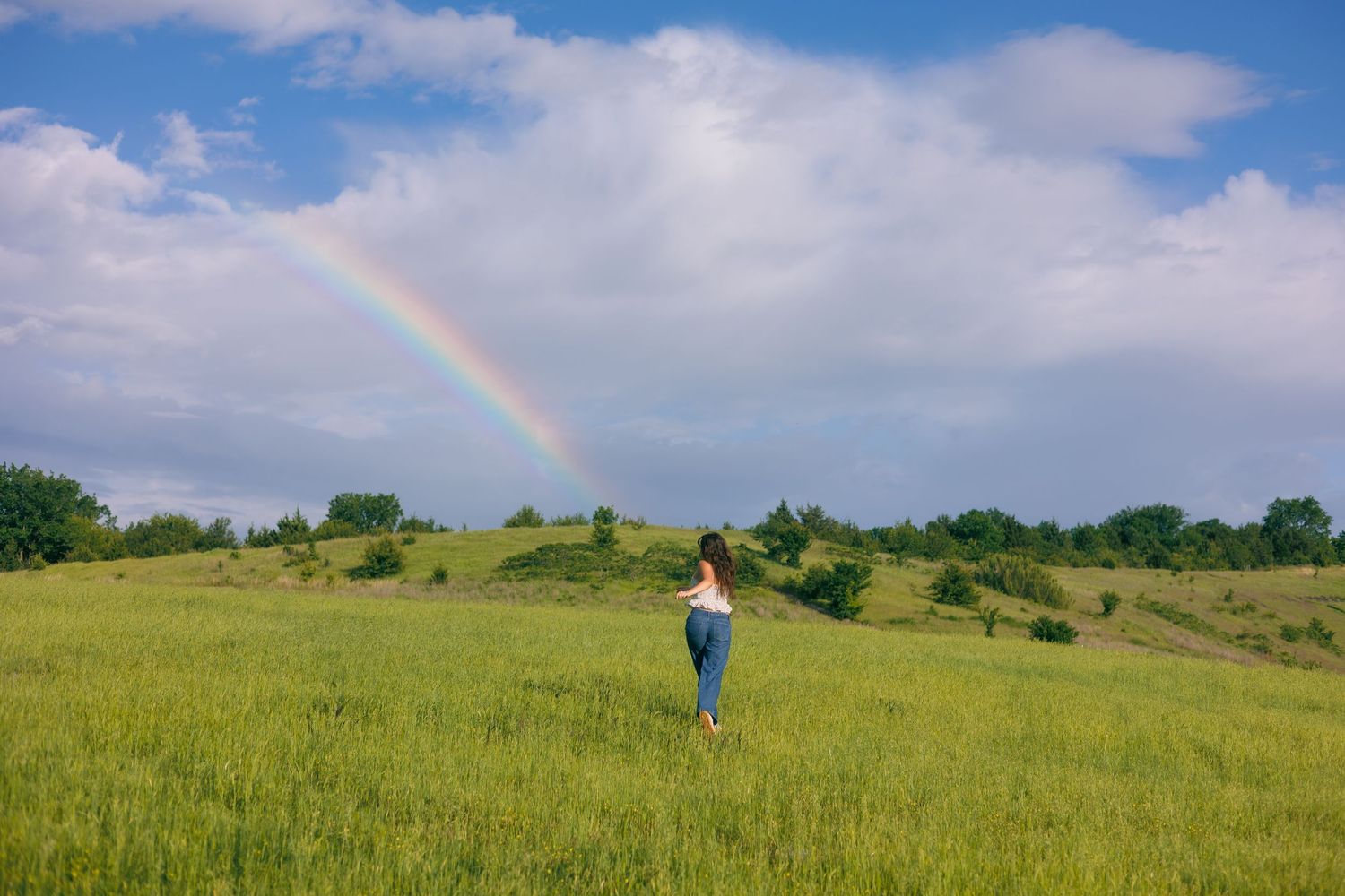Someone stands in a vast green field while a rainbow curves through clouds above.